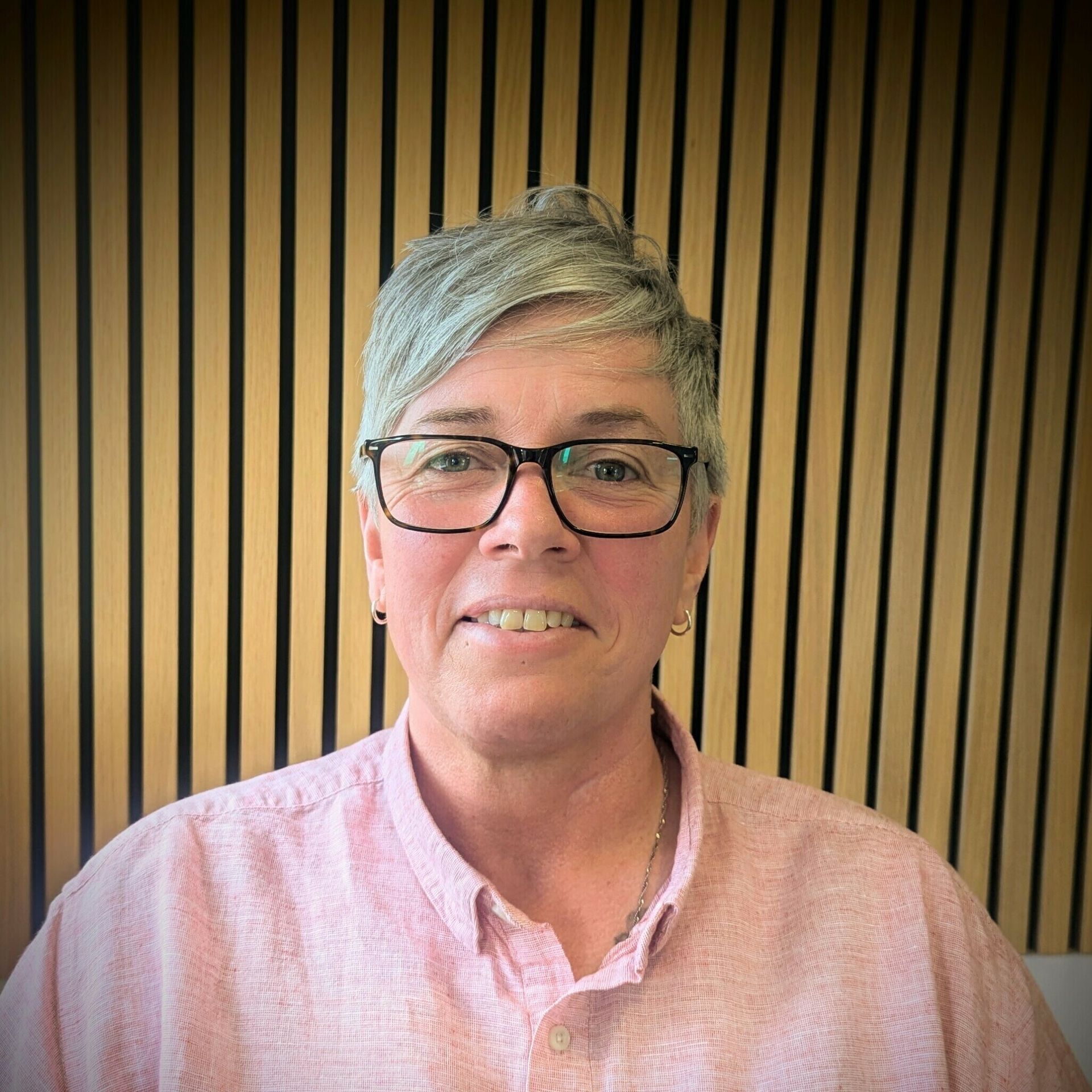 Person wearing glasses and a pink shirt, smiling. Background is wooden panels.