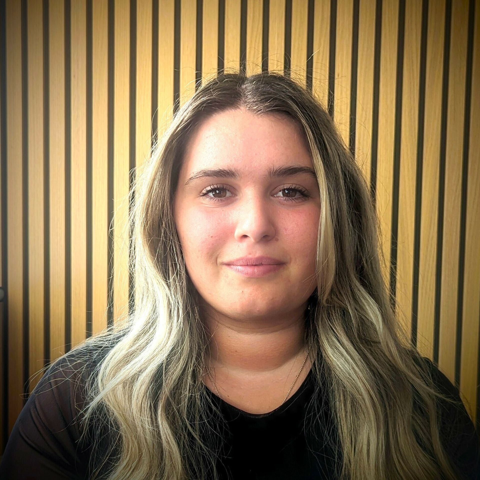 Woman with wavy, light-colored hair smiles at camera, wearing a black shirt, against a wooden panel background.