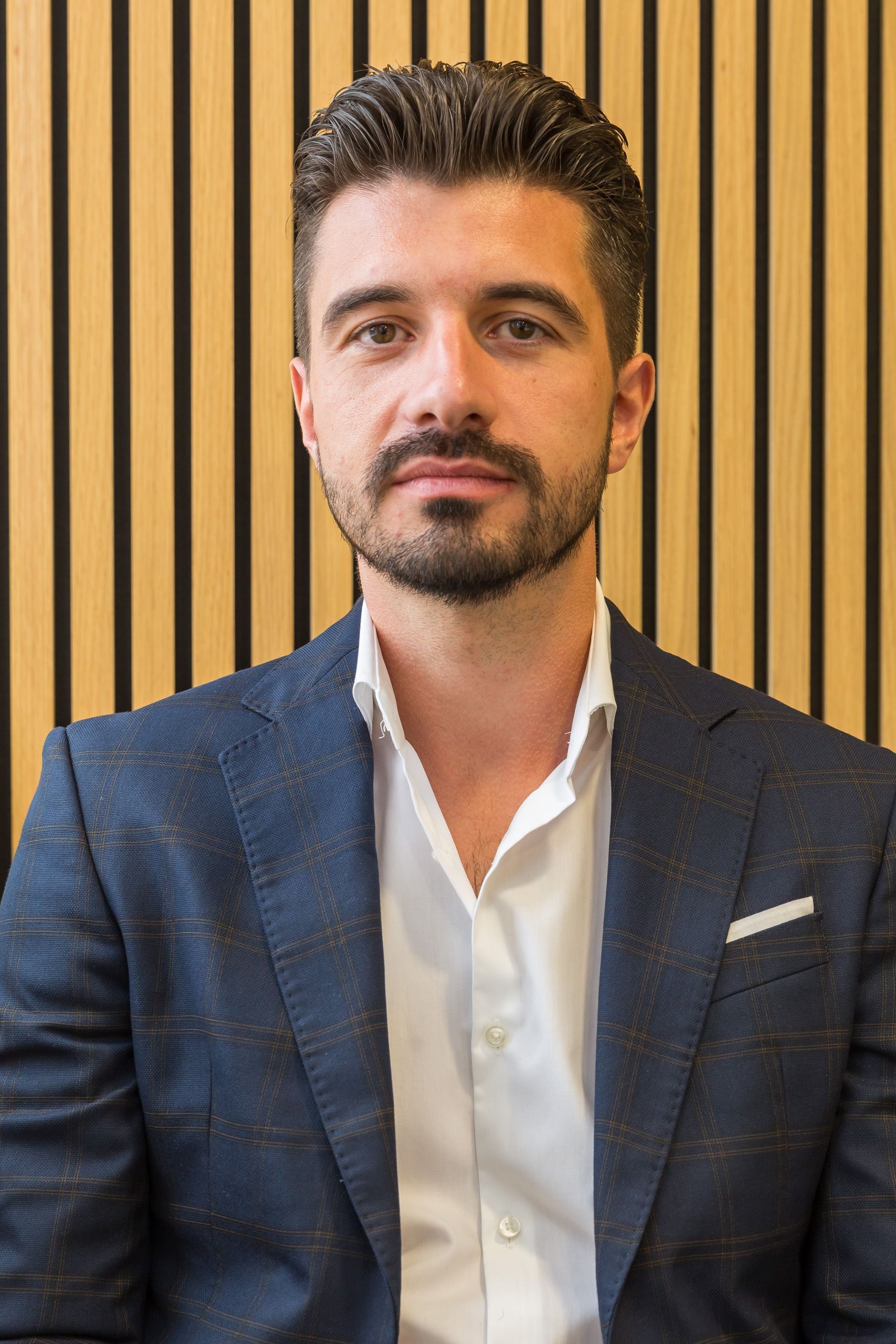 Man in a blue blazer and white shirt, looking at the camera in front of a wood-paneled wall.
