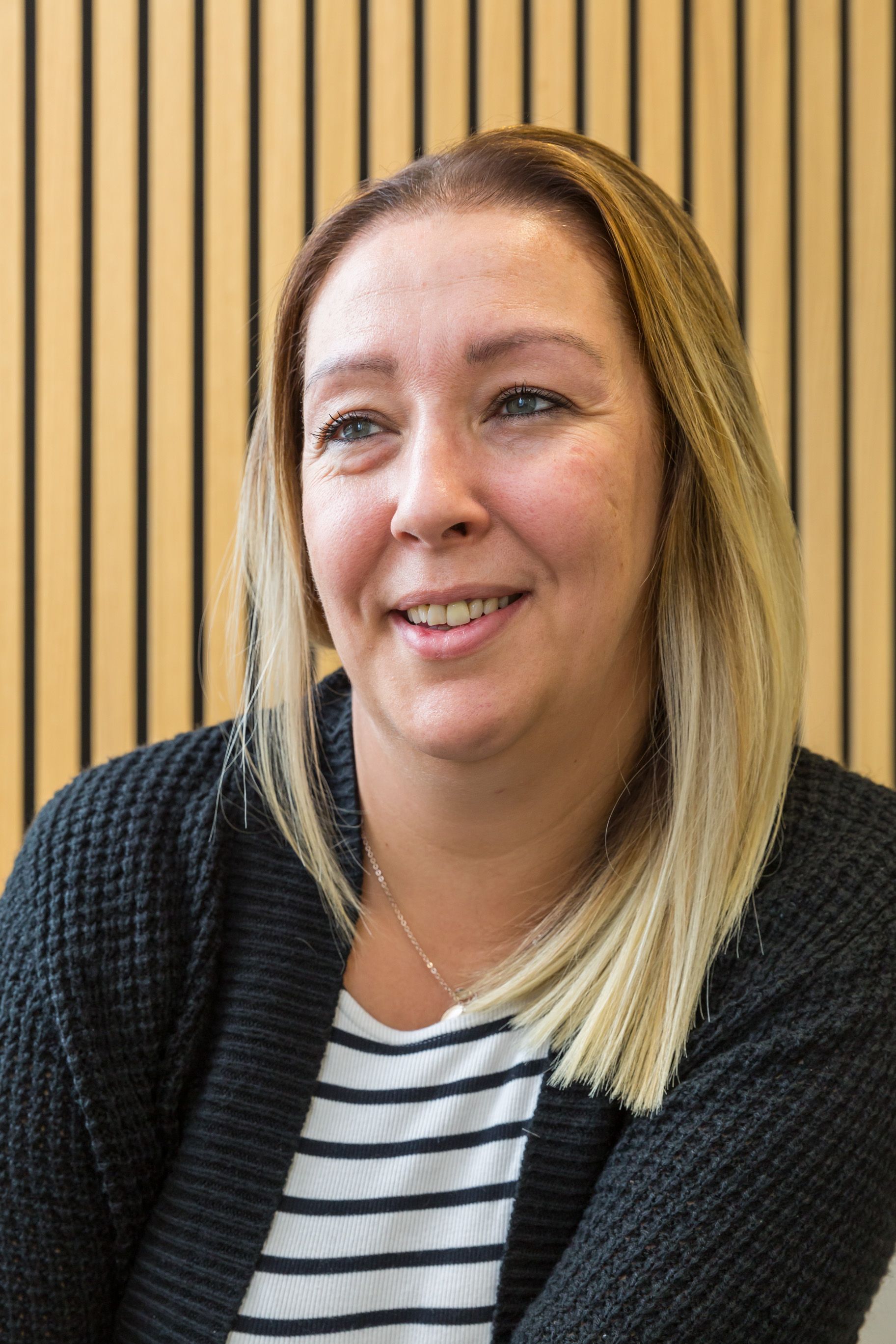 Woman with blonde-streaked hair smiles, wearing a black sweater over a striped shirt, seated in front of a wooden wall.