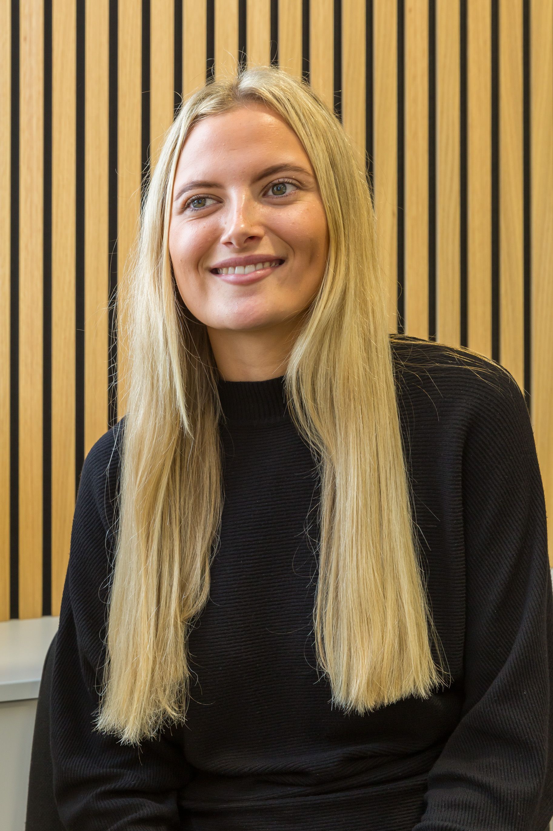 Blonde woman with long hair smiles, wearing black sweater, in front of wood panel wall.
