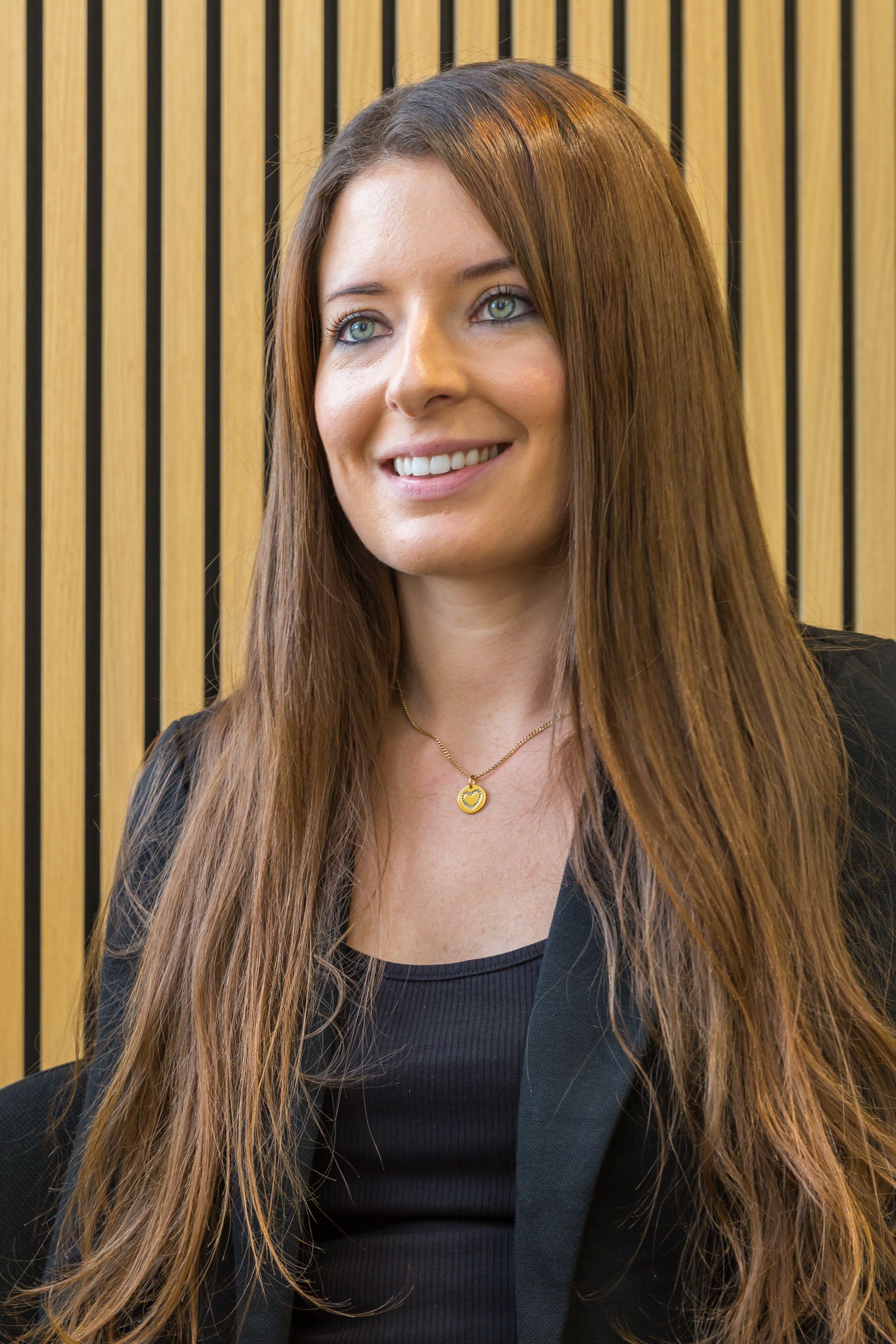Woman with long brown hair smiles, wearing a black blazer and top, in a wood-paneled room.