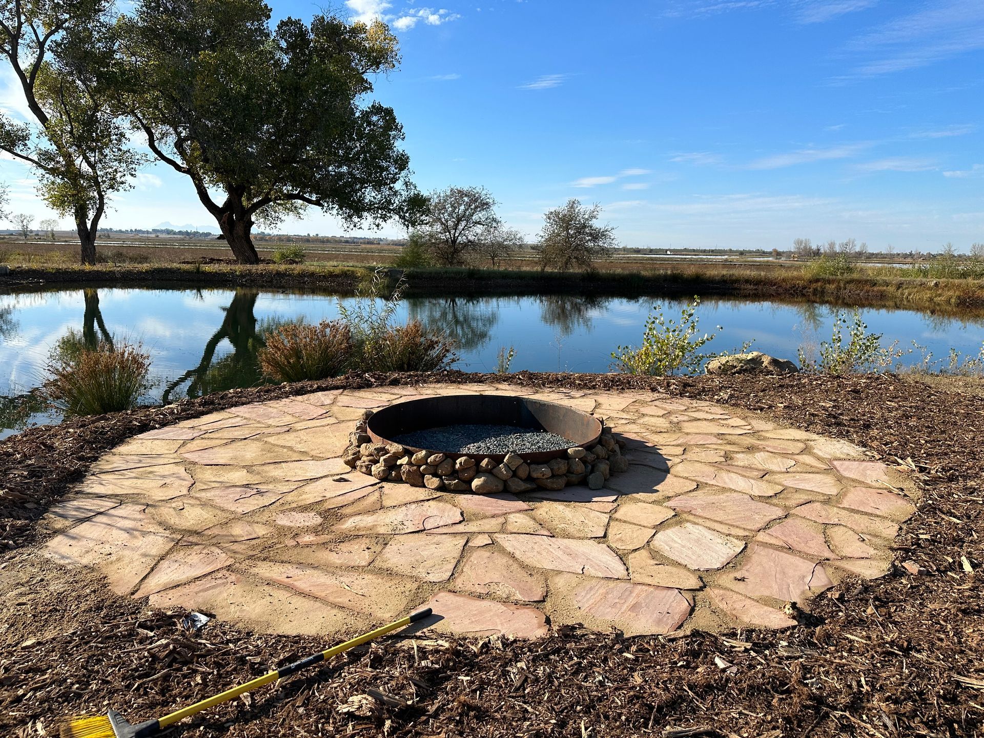 A fire pit is sitting in the middle of a stone patio next to a pond.