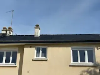 A light-colored house exterior with three white-framed windows, a central chimney, and a dark shingled roof under a blue sky.