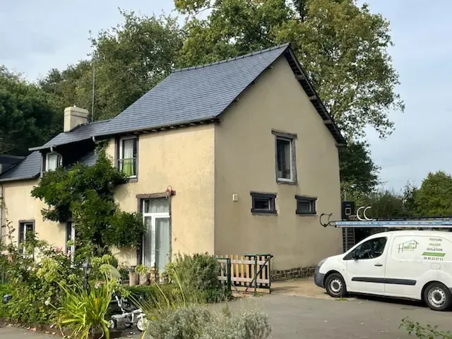 A light-colored two-story cottage with a dark slate roof, surrounded by trees and a garden, with a white van parked nearby.