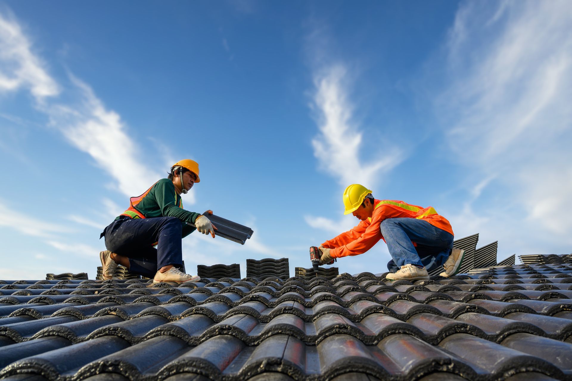 Two workers in hard hats and safety vests install dark roof tiles against a bright blue sky.