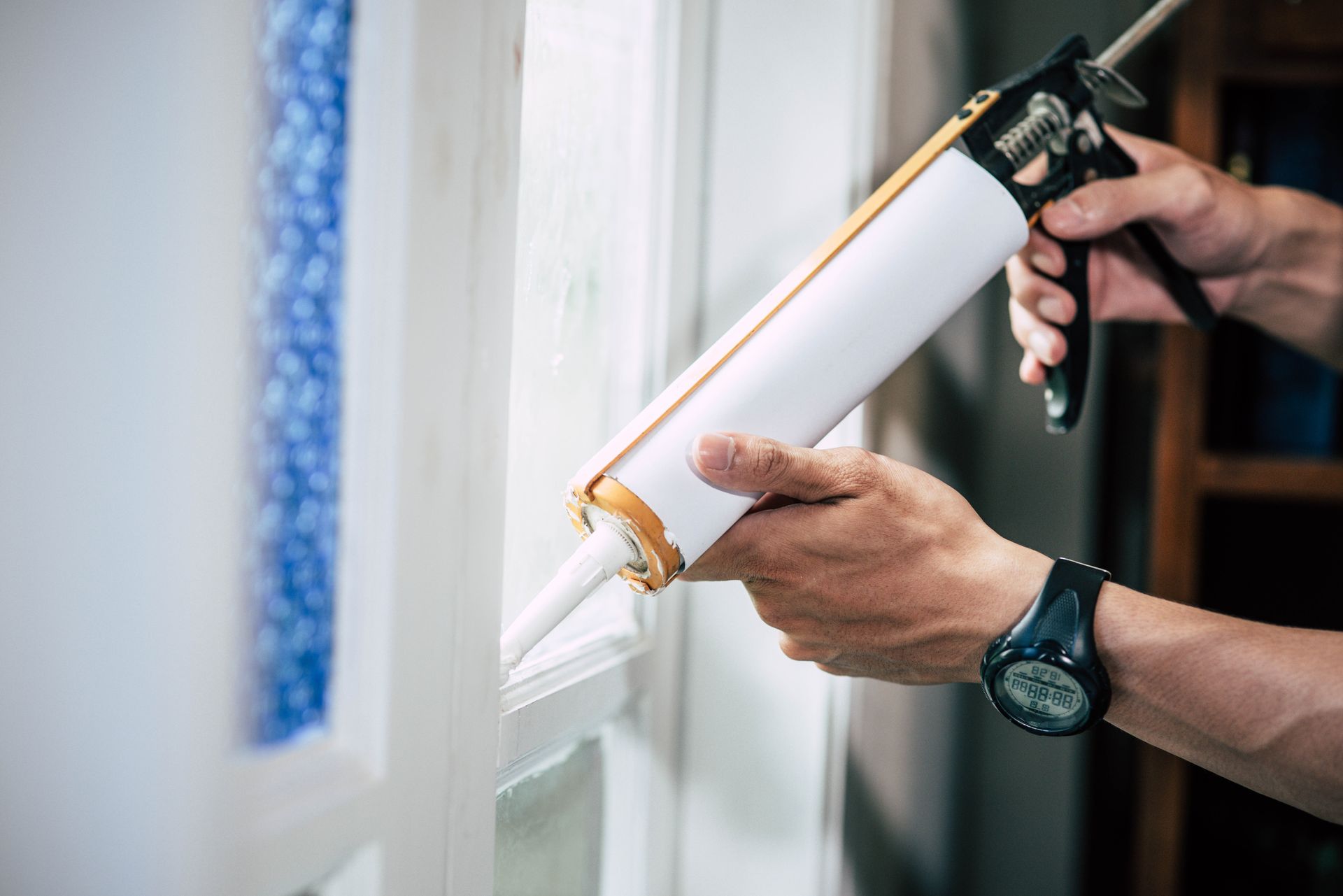 A person uses a caulk gun to apply sealant along the edge of a white window frame.