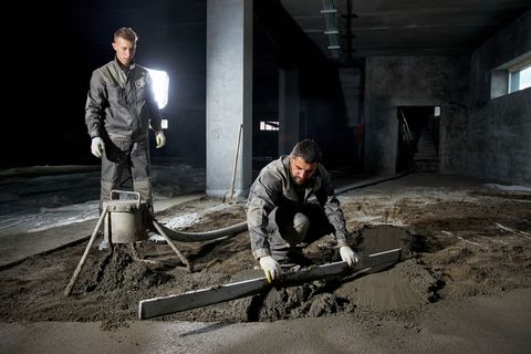 Two workers in grey uniforms smooth wet concrete on an industrial building floor using a long metal leveling tool.