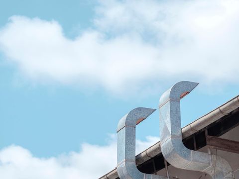 Two galvanized metal ventilation ducts extending from the edge of a building roof against a bright blue sky with clouds.
