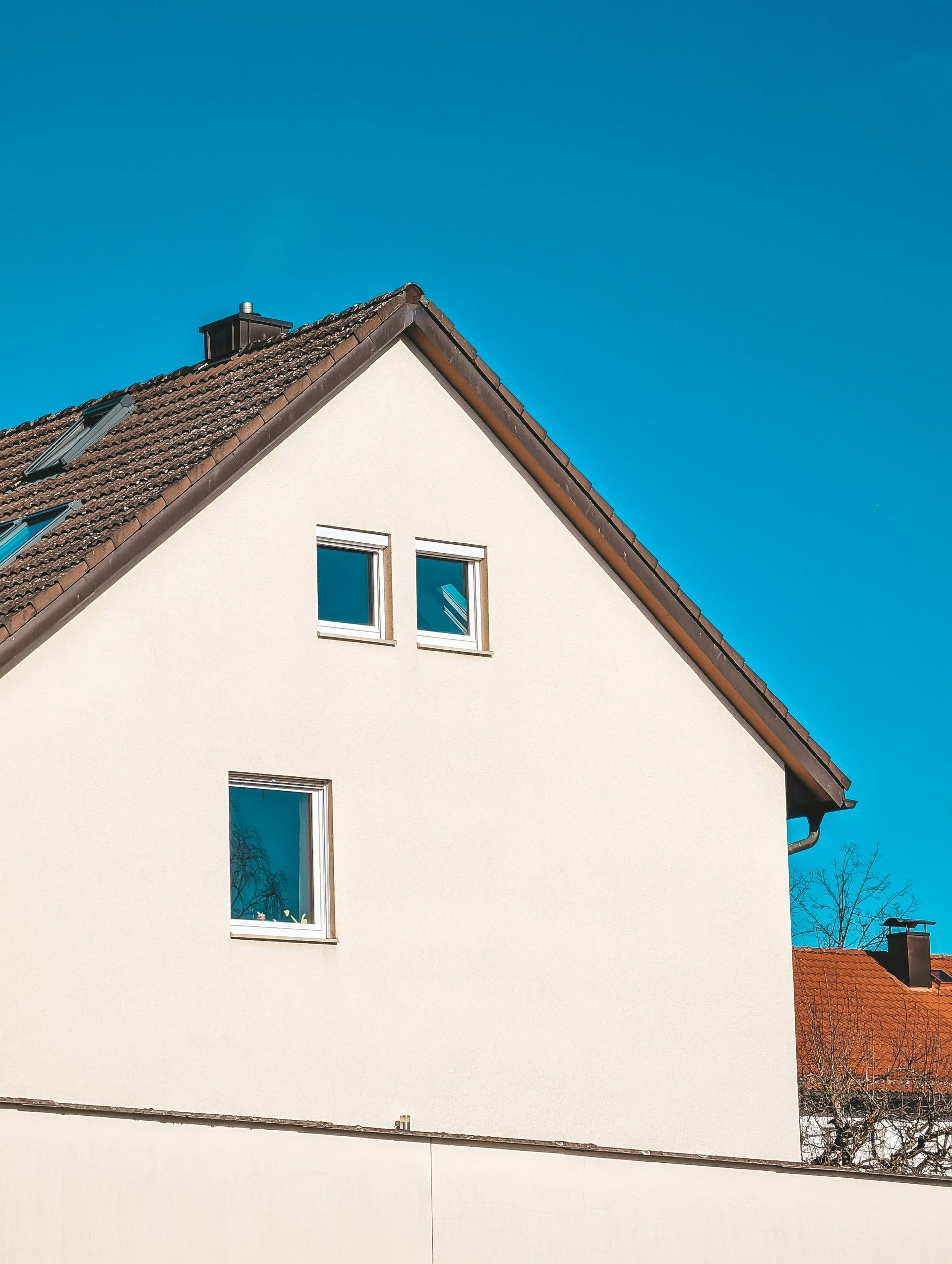 A light-colored house with a dark tiled roof against a clear blue sky, featuring three windows on the side gable.