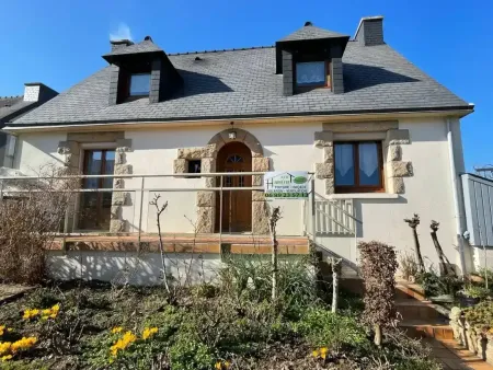 A dormer window with black trim and white curtains on a roof covered in dark shingles next to a white brick chimney.