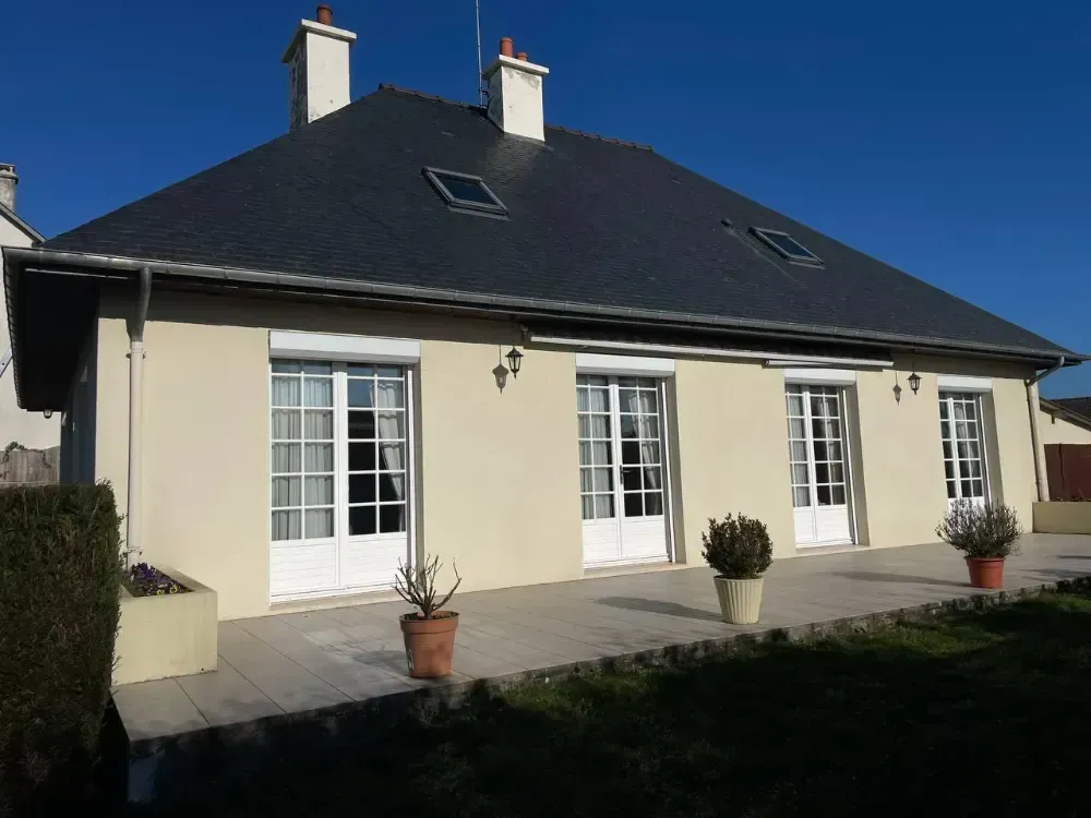 A beige house with a dark slate roof, four white French doors, two chimneys, and a stone patio under a clear blue sky.