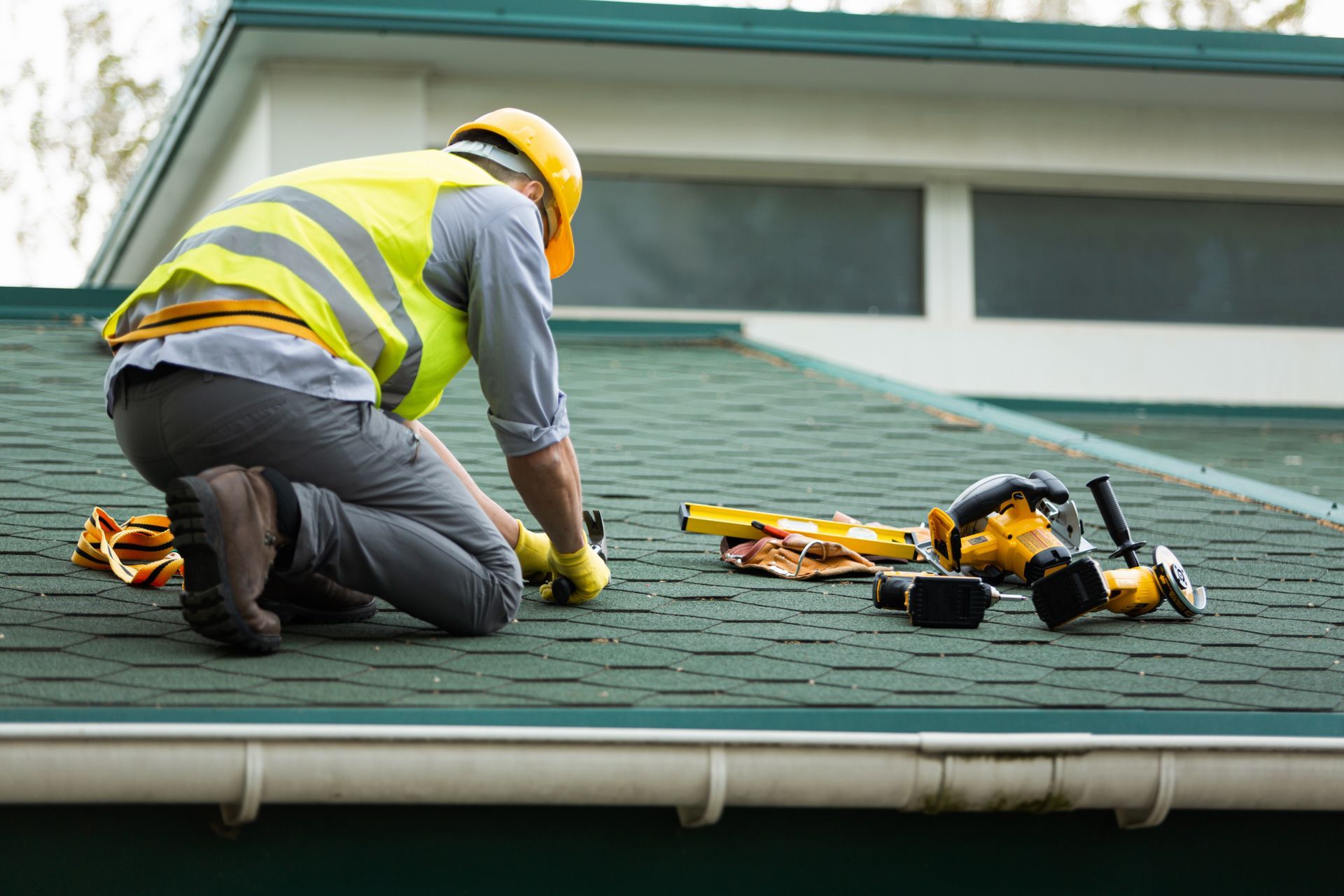 A worker in a yellow high-visibility vest and hard hat kneels on a green roof, installing shingles with tools nearby.