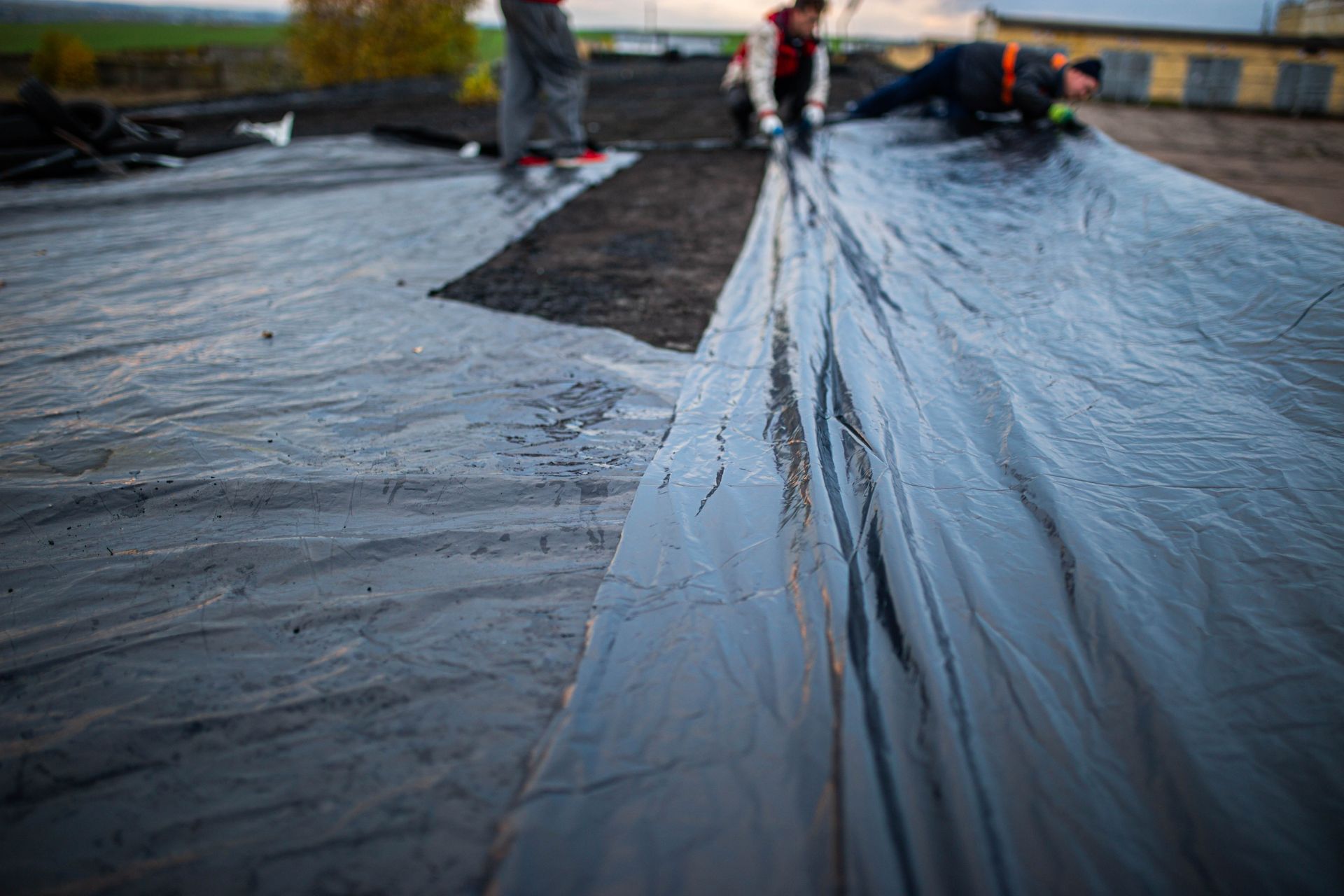 Construction workers unroll thick, black waterproof sheeting across a flat rooftop during a construction project.