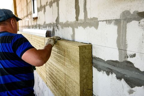 A person in a blue striped shirt installs insulation boards onto a white block wall.