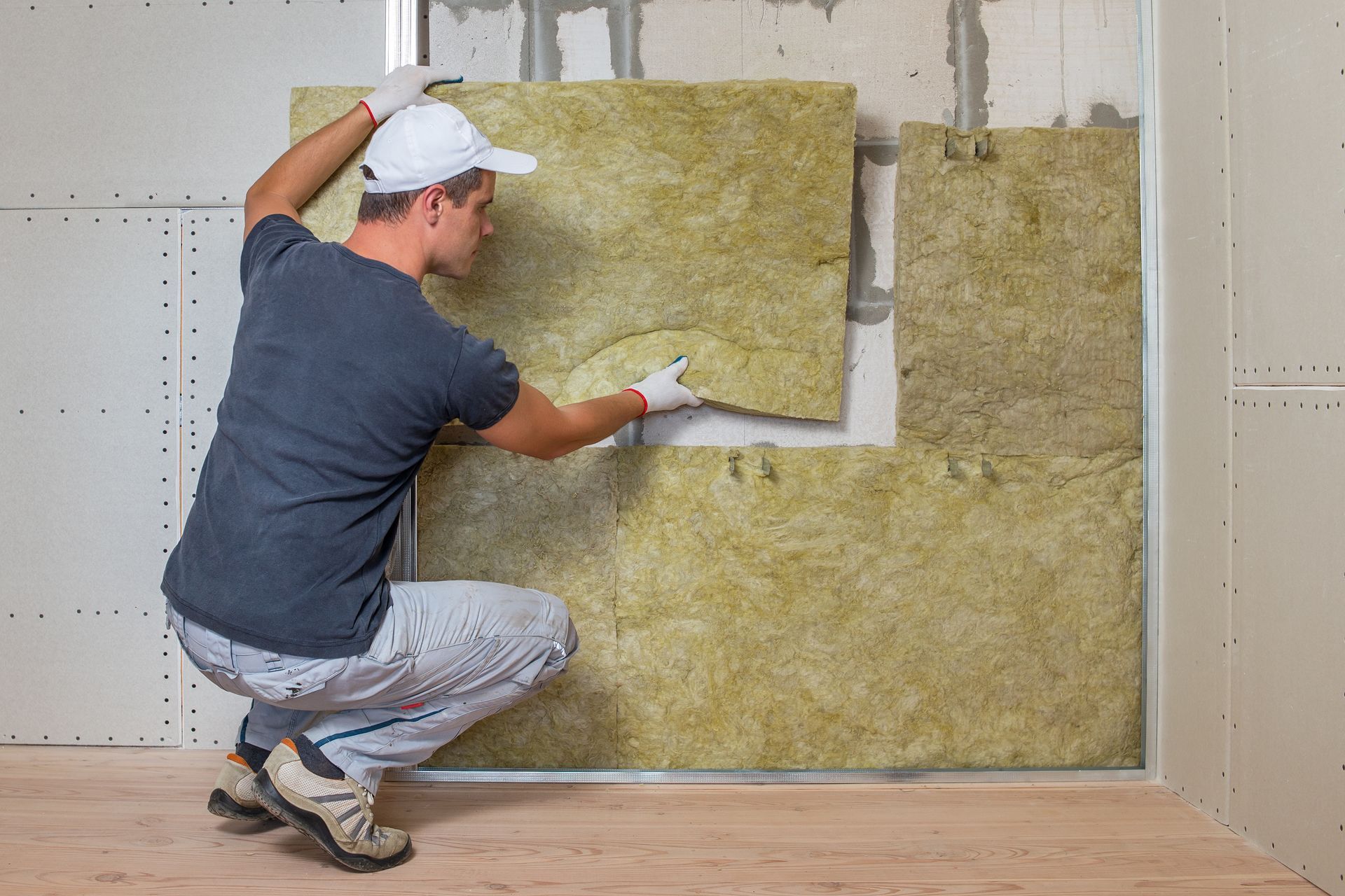 A worker in a white cap and gray clothing installing yellow insulation panels onto an interior wall.
