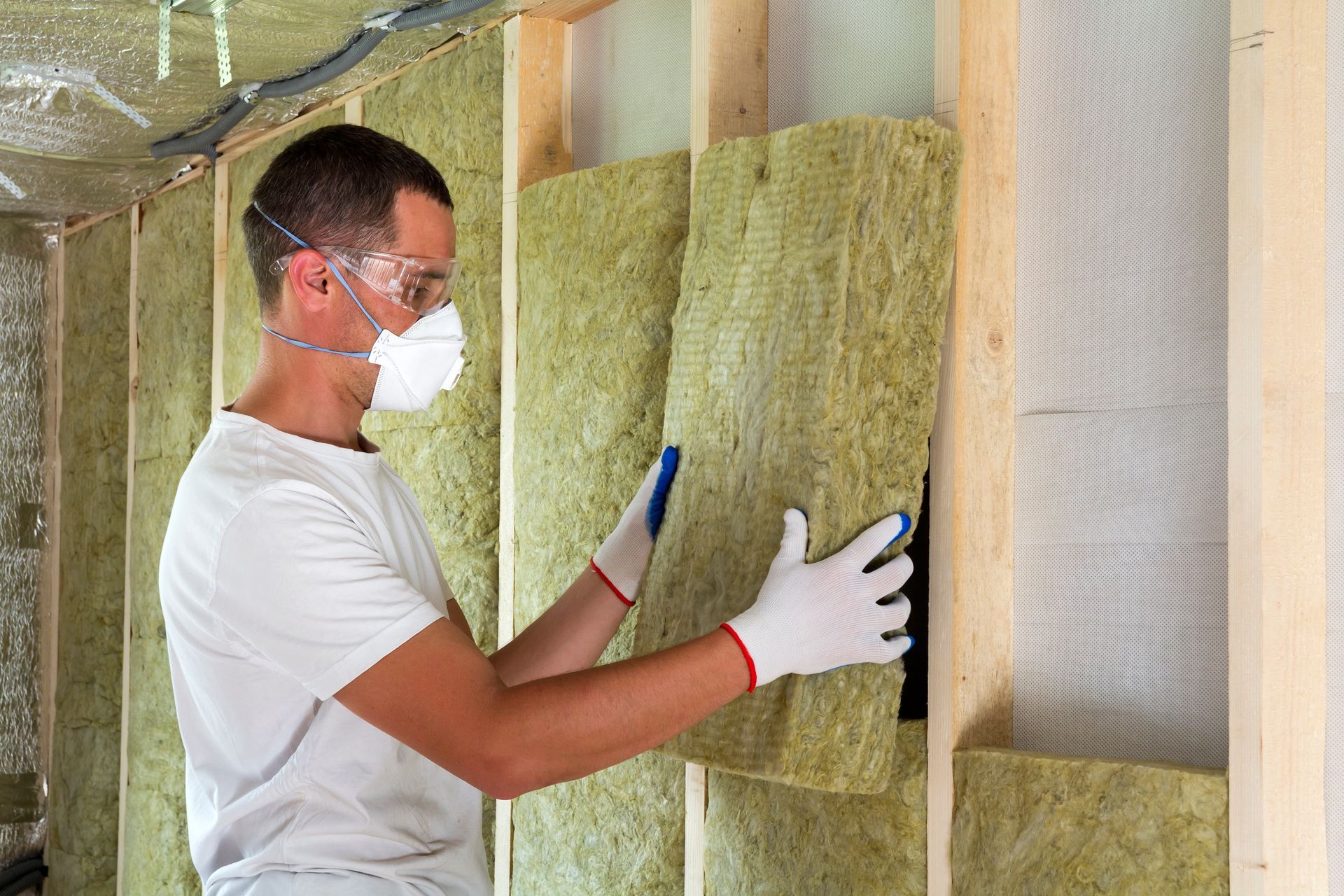 A person wearing protective eyewear, a mask, and gloves installs mineral wool insulation between wooden wall studs.