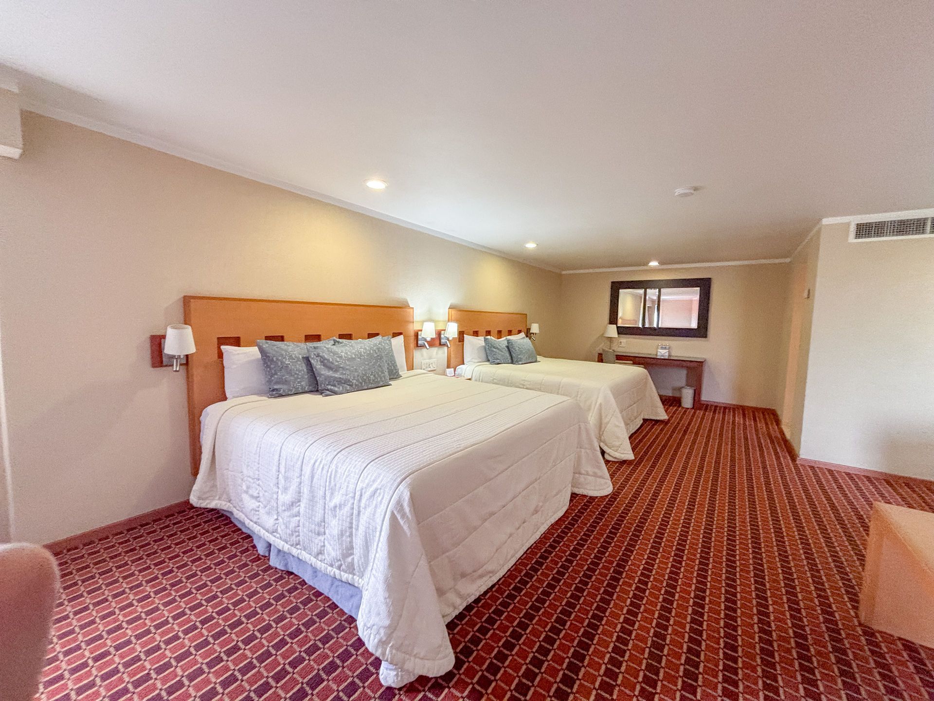 Una habitación de hotel con dos camas blancas tamaño queen, un escritorio, una ventana y una alfombra roja estampada.