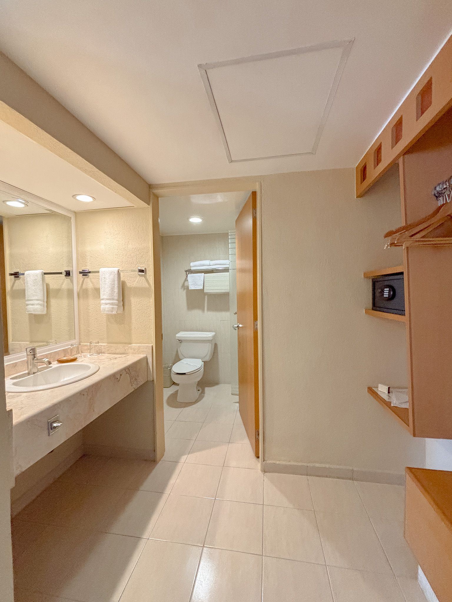 Un baño de hotel con tocador, un armario abierto con estantes y caja fuerte, y una habitación separada con inodoro.