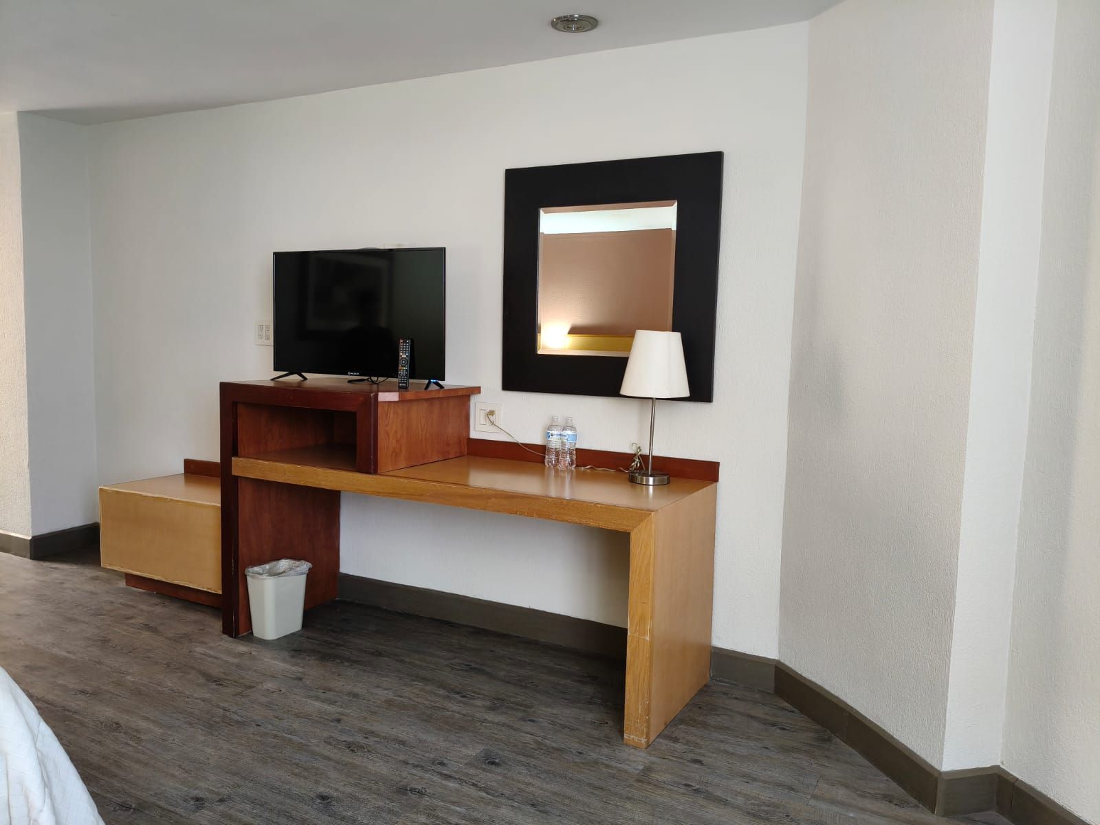 Un escritorio de madera con un televisor de pantalla plana, un espejo cuadrado y una pequeña lámpara 