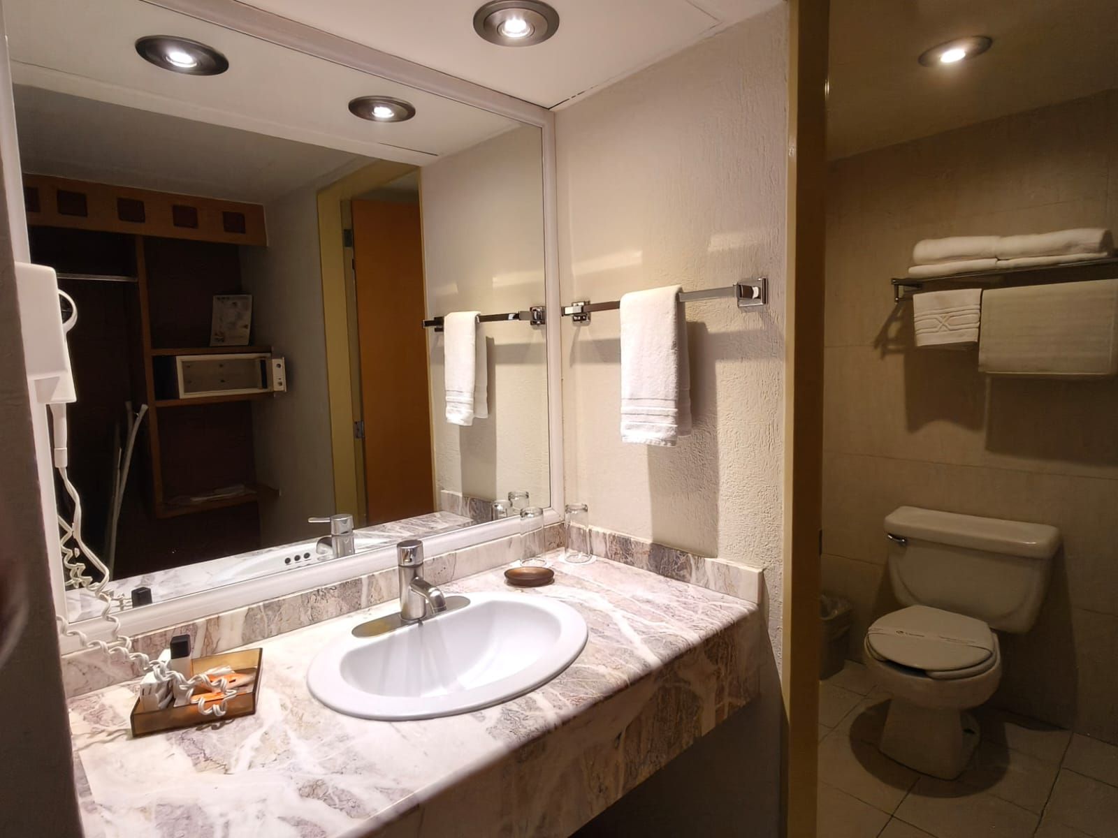 Un baño de hotel con tocador de mármol con lavabo, un espejo grande y un área de inodoro contigua con toalleros.