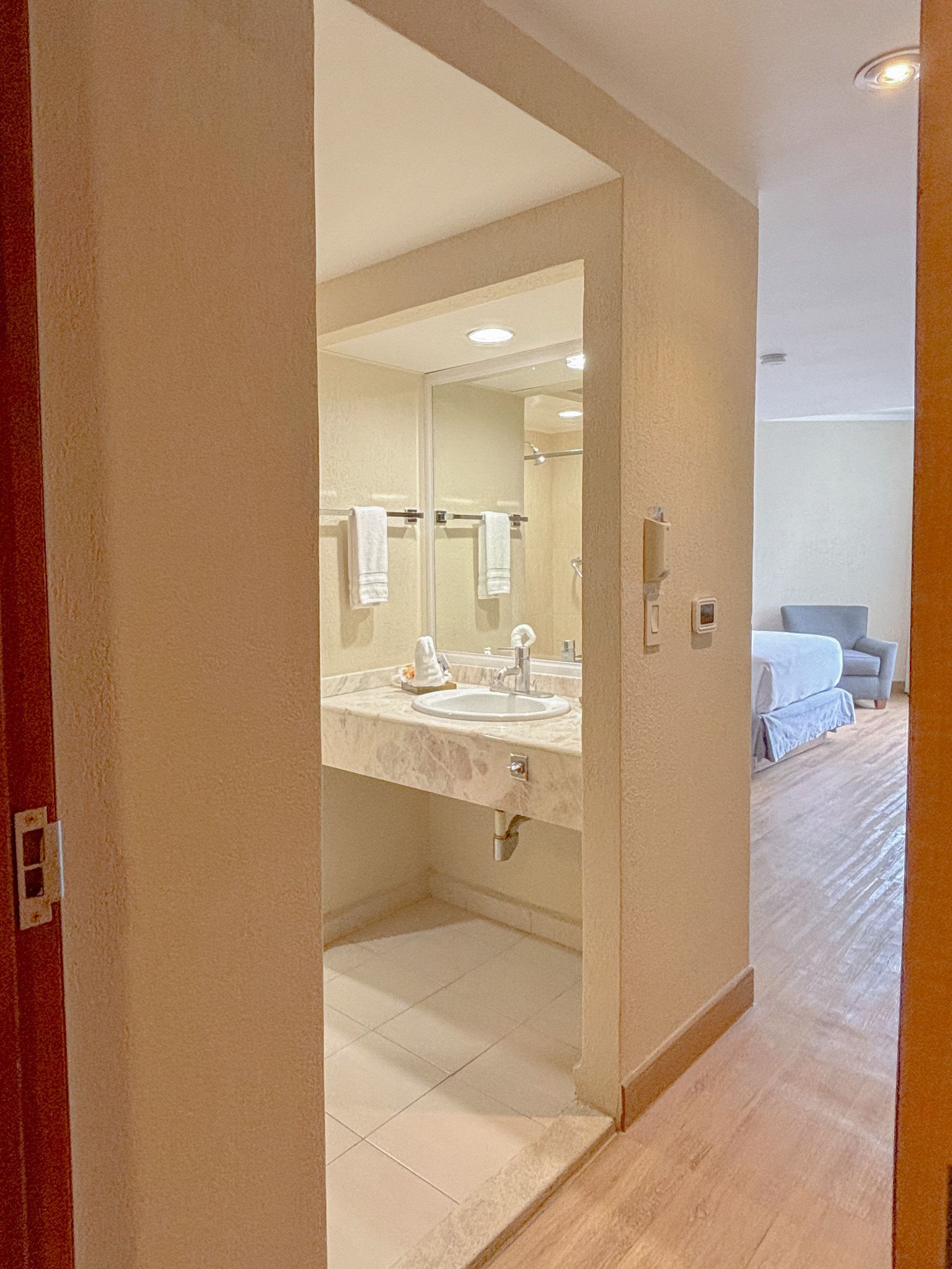 Se ve un tocador de baño con espejo y encimera blanca a través de una puerta, con un dormitorio al fondo.