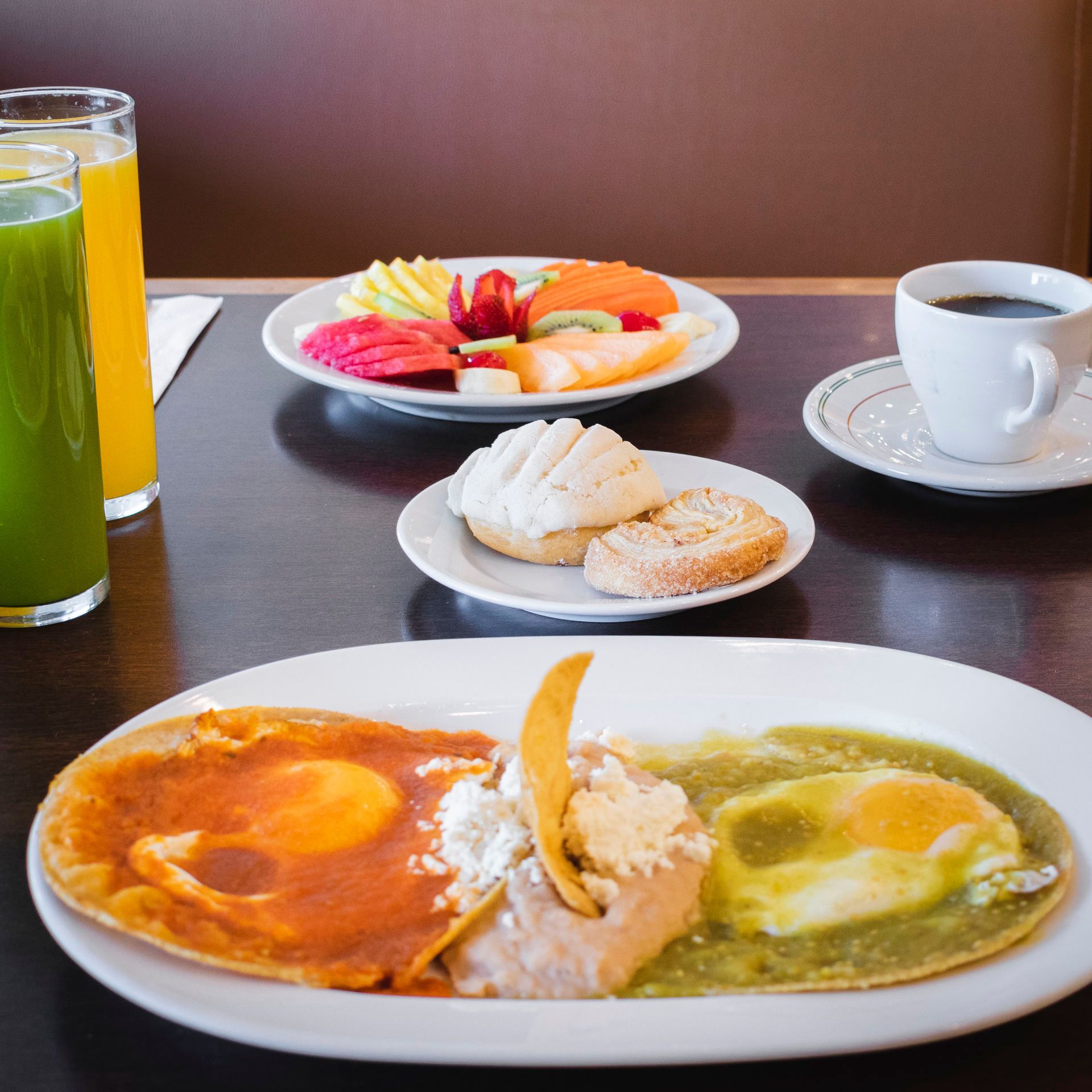 Un desayuno completo que incluye huevos divorciados, rodajas de fruta, bollería, una taza de café y dos vasos de zumo.