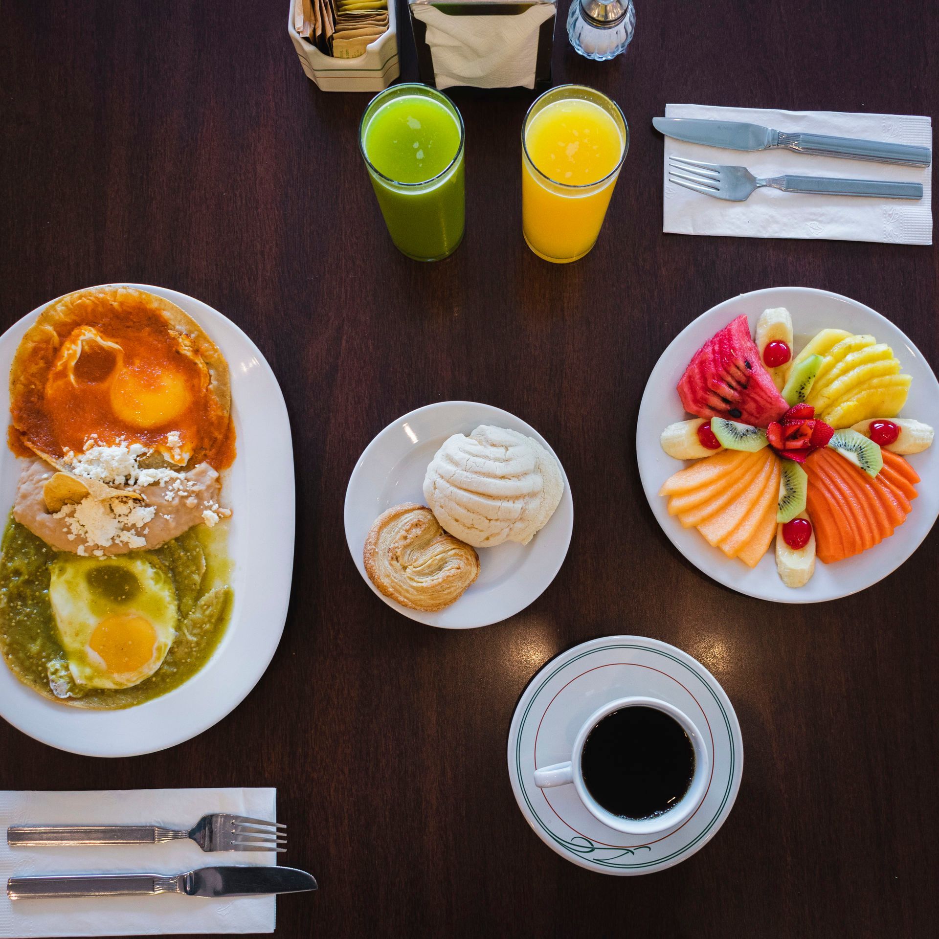 Vista desde arriba de un desayuno servido sobre una mesa oscura, con huevos, fruta fresca, pan dulce, zumo y café.