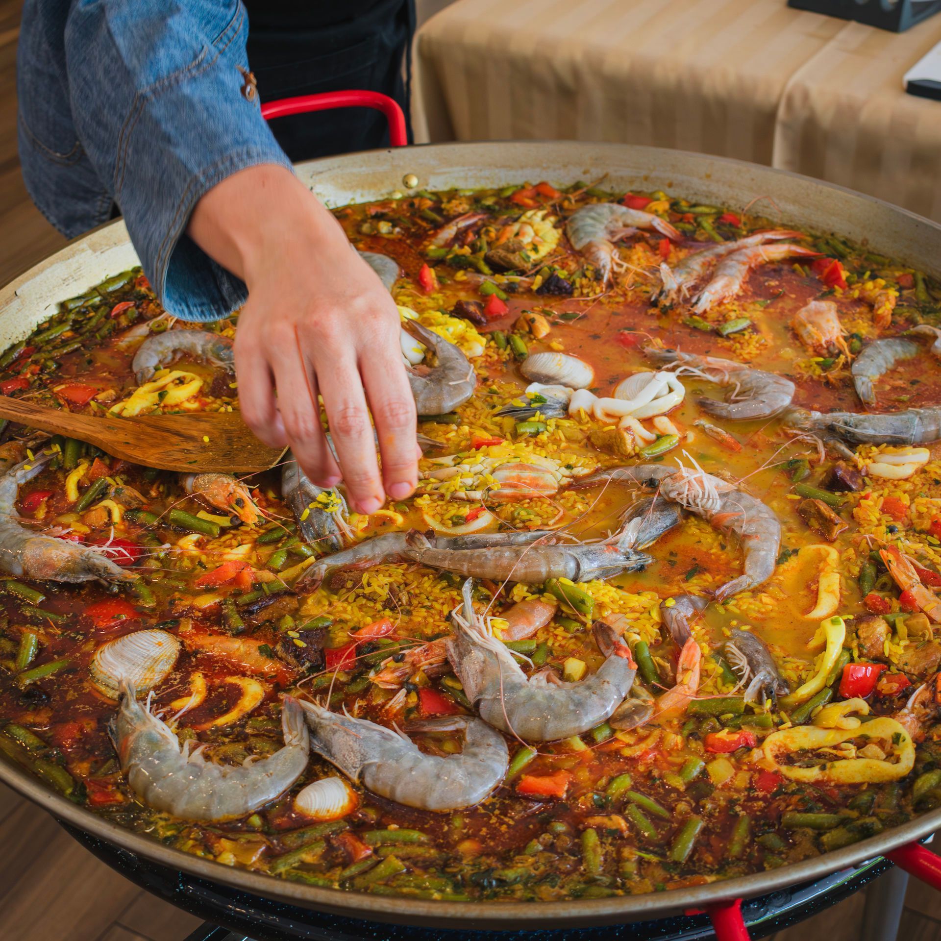 Una persona prepara una paella de marisco grande con gambas y verduras, añadiéndoles condimentos a mano.