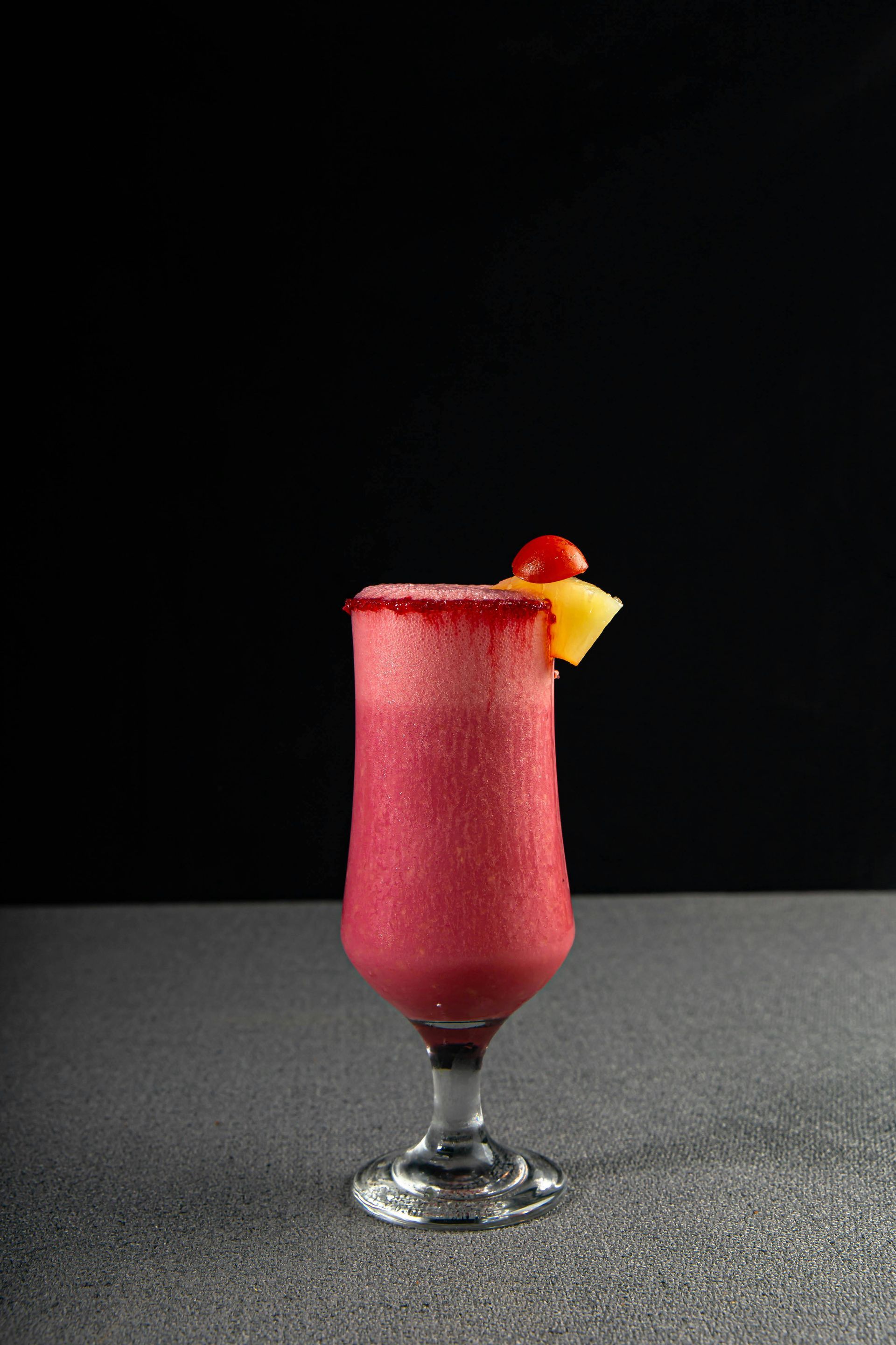 Un cóctel rojo helado servido en una copa con tallo y borde azucarado, adornado con una rodaja de piña y una cereza.