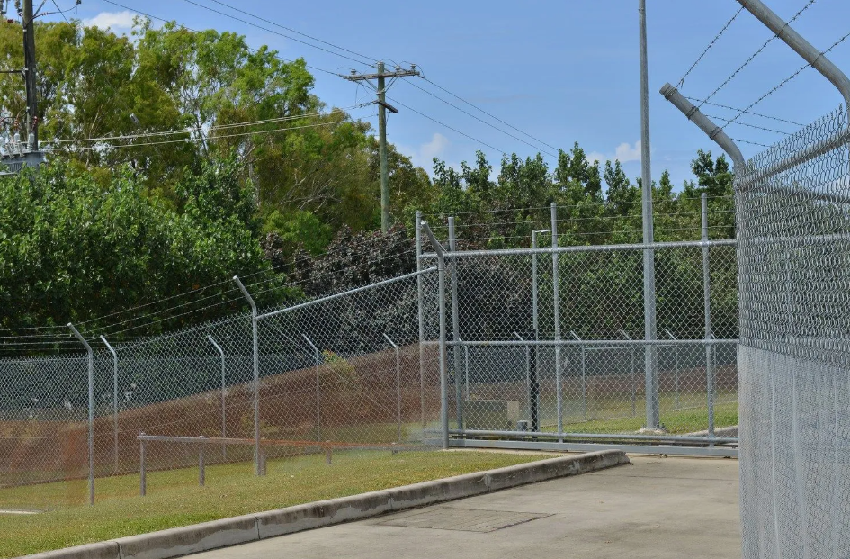 Chain-link fence topped with barbed wire surrounds a facility with a gate; trees and power lines in the background.