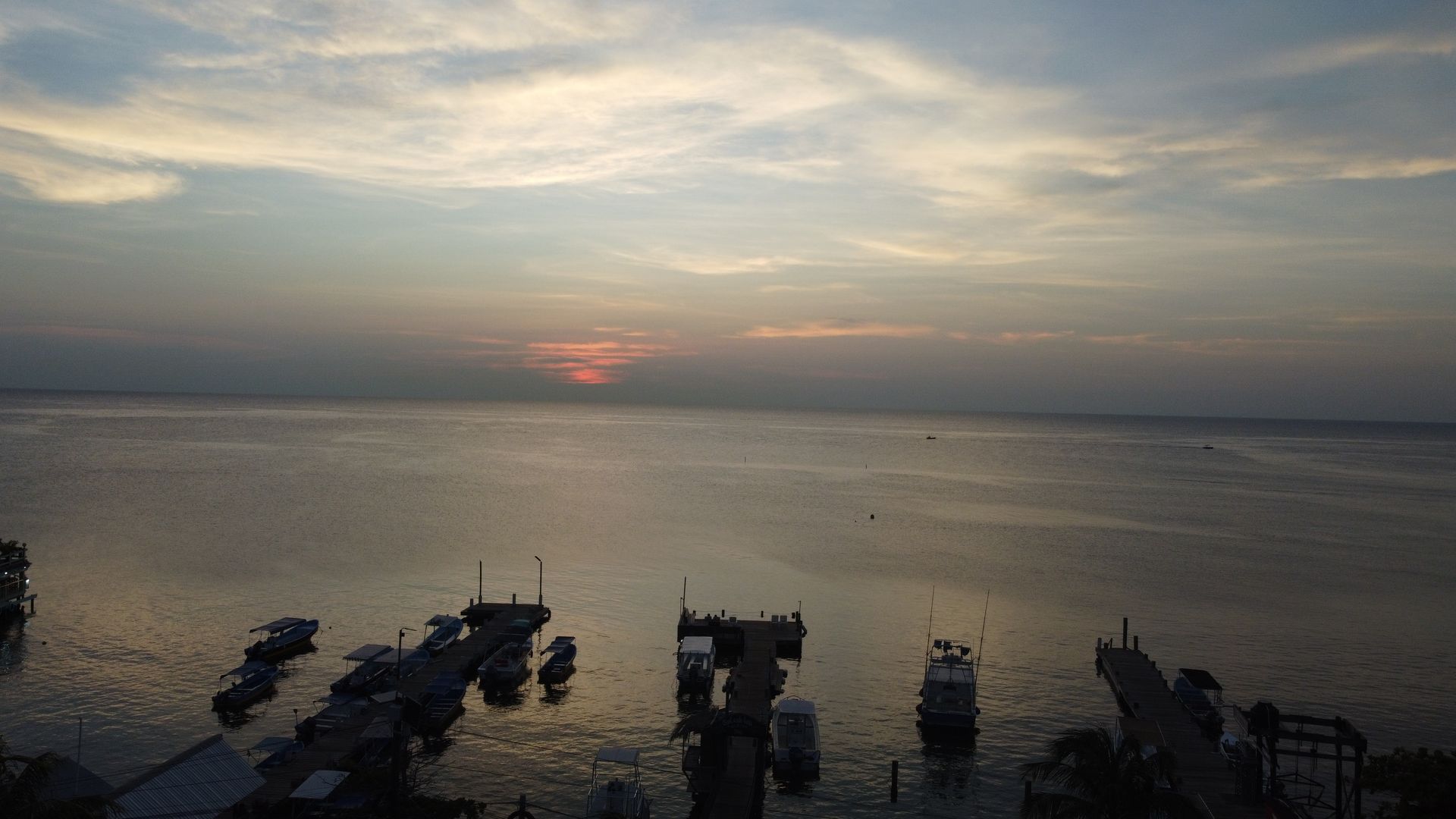 Sunset over a body of water, silhouetted boats at a dock. Pale blue and orange sky.