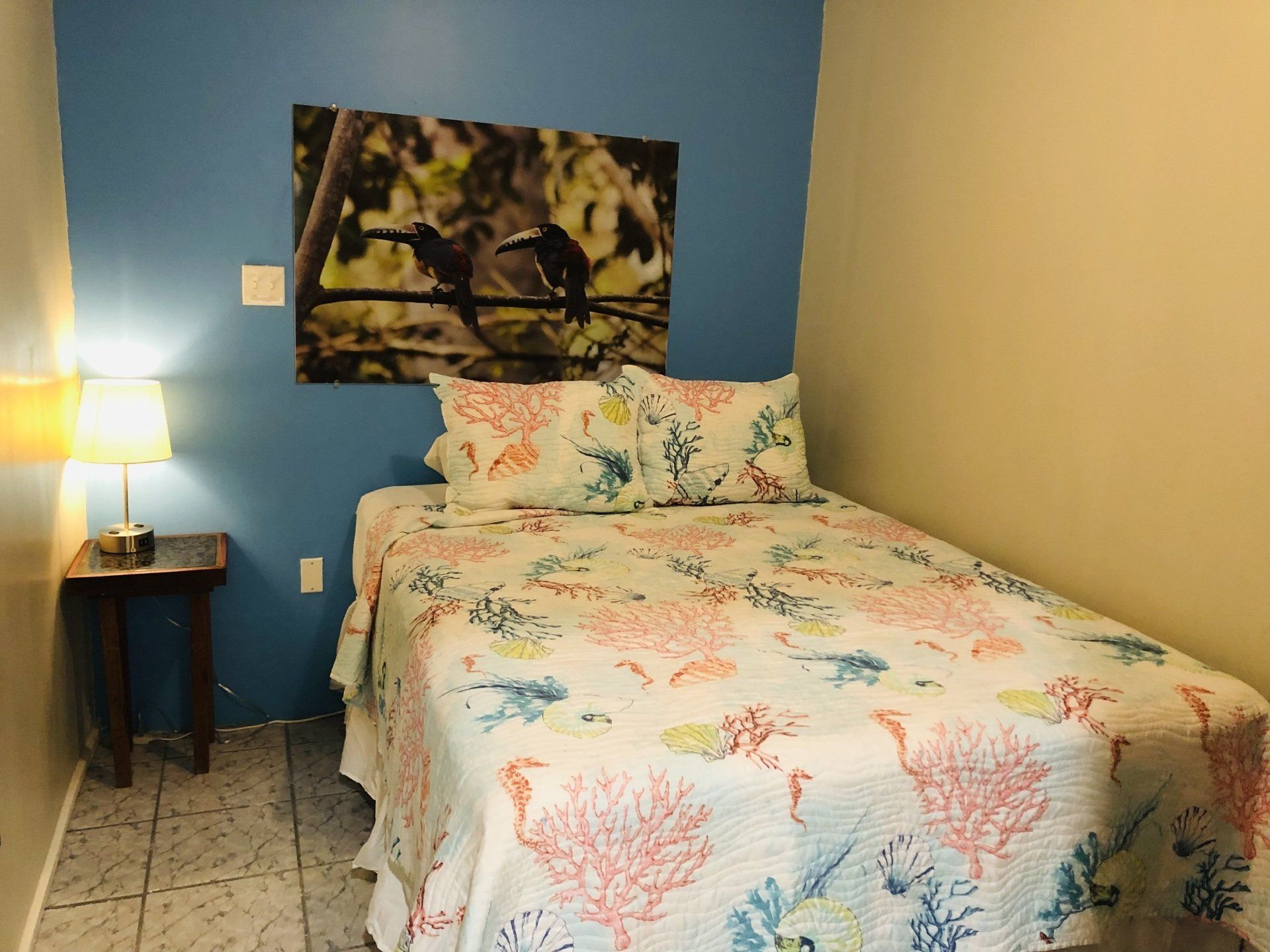 Bedroom with a single bed, coral-themed bedding, toucan art, blue and beige walls, lamp on a nightstand.