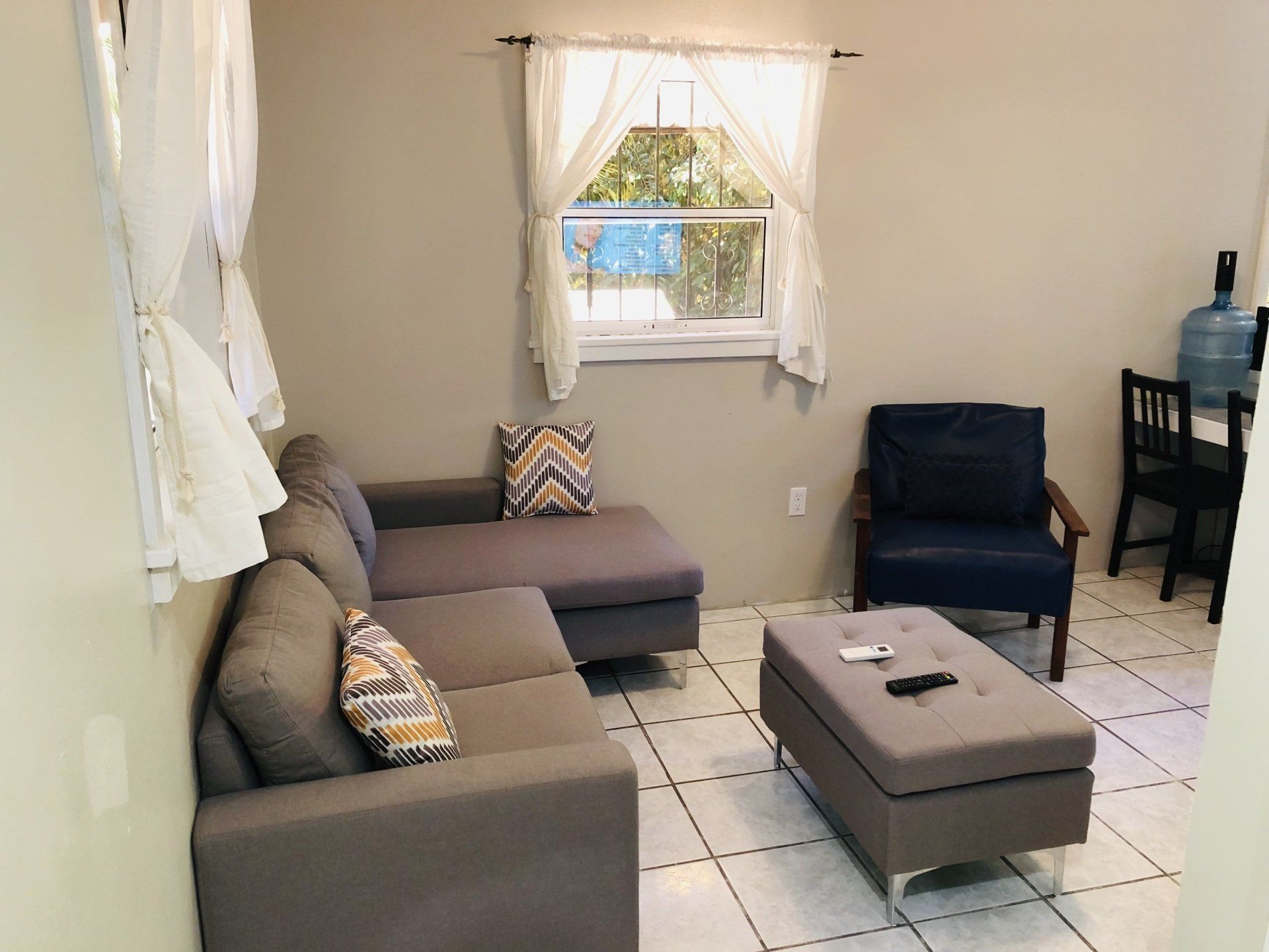 A living room with gray sectional sofa, blue armchair, window with white curtains, and a water jug.