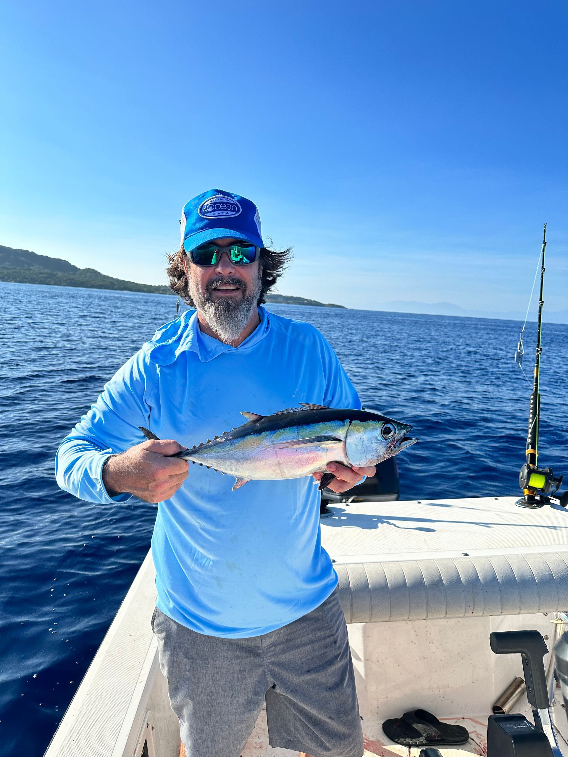 Man on a boat holding a fish. Blue ocean and sky. He wears a blue shirt, hat, and sunglasses.