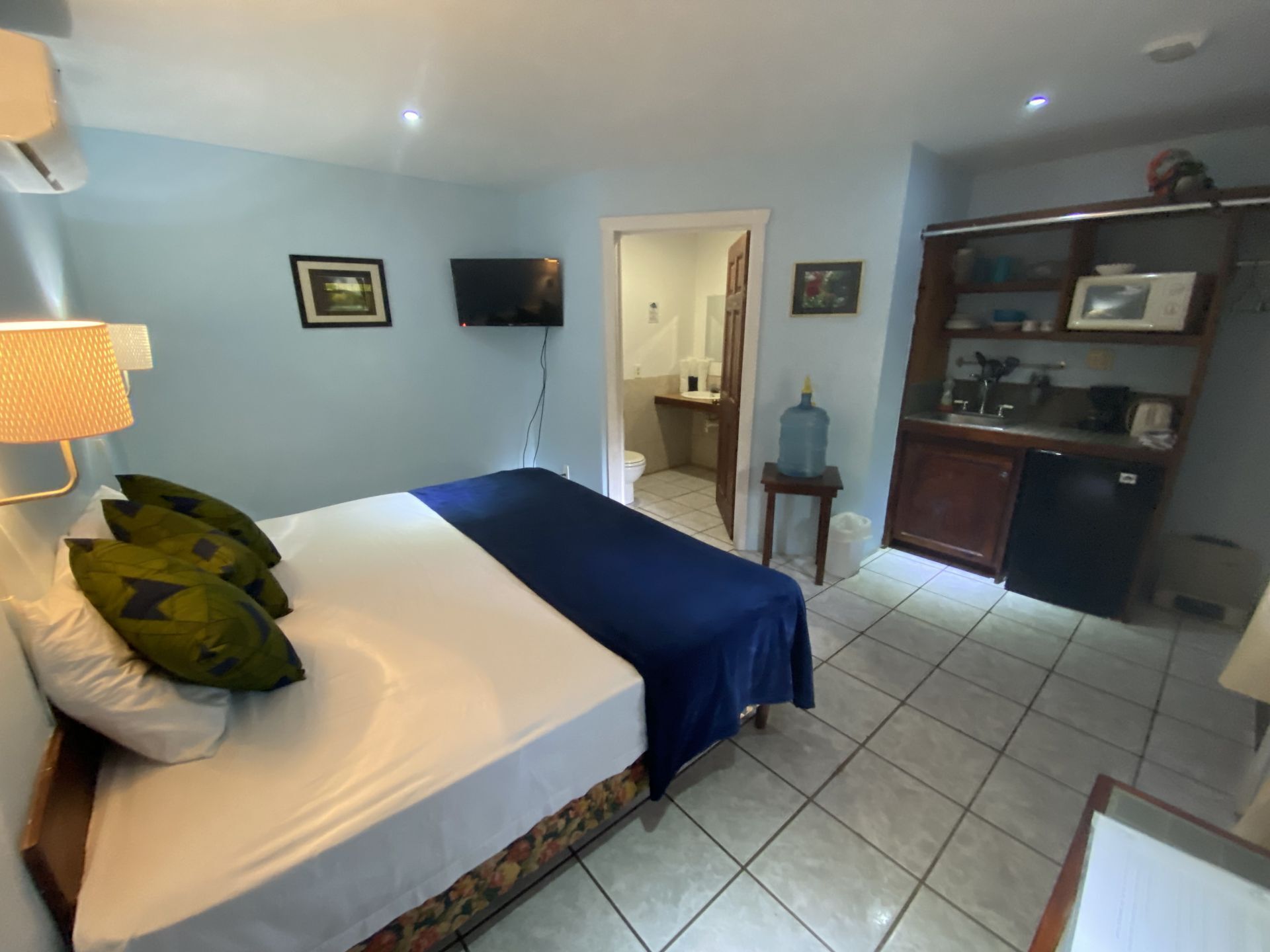 Blue-walled hotel room with bed, TV, small fridge, and bathroom visible through doorway.