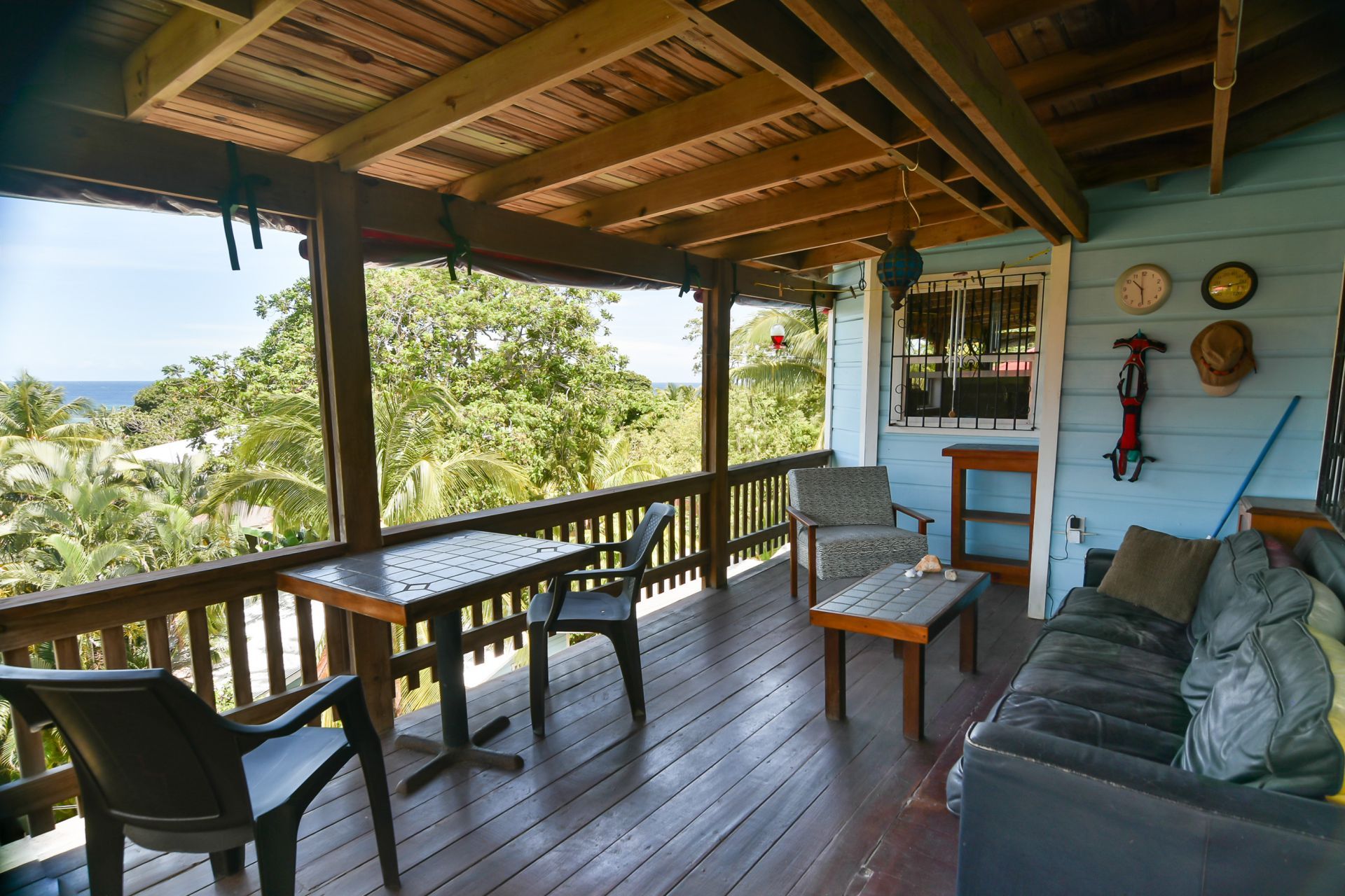 Wooden porch with table, chairs, and couch overlooking lush green trees and the sea.