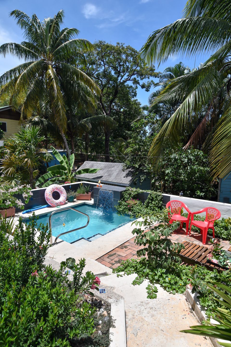Small pool surrounded by lush greenery, red chairs, and a palm tree under a sunny sky.