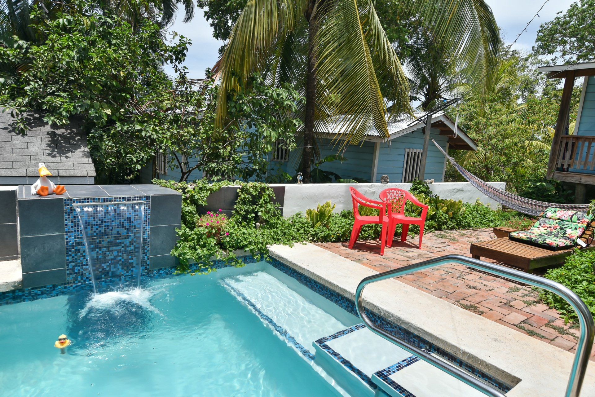 Pool with waterfall, red chair, hammock, and small buildings surrounded by greenery.
