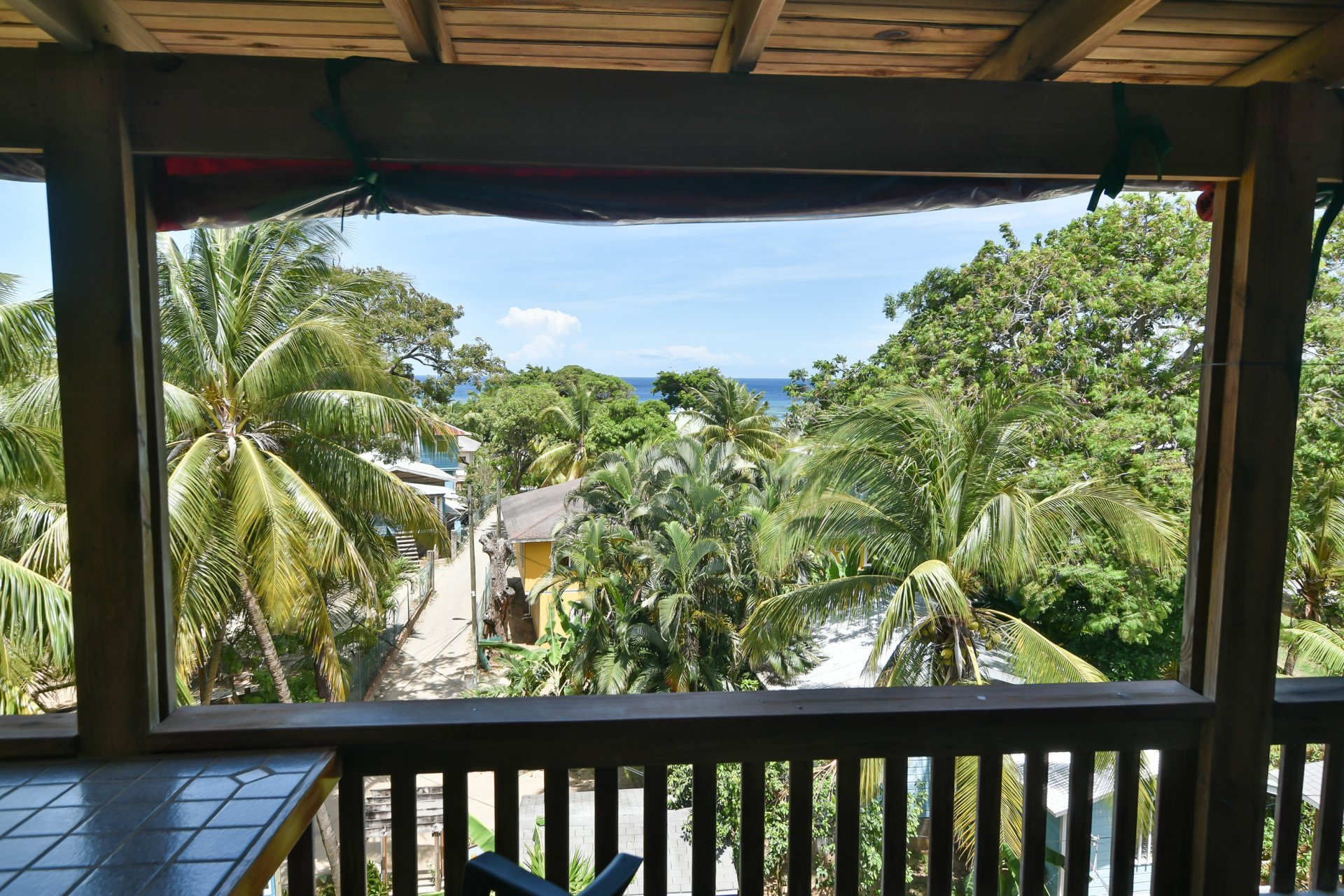 View of a lush tropical scene through a wooden porch: palm trees, blue sea, and sky.