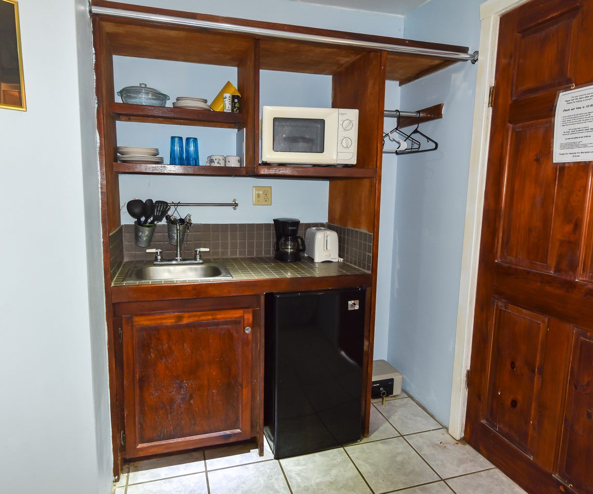 Small kitchenette with a microwave, sink, fridge, and shelves next to a wooden door.