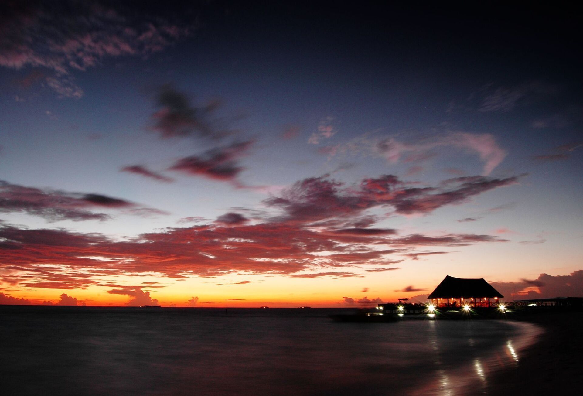 Sunset over the ocean, with a silhouetted hut and vibrant orange and purple clouds.