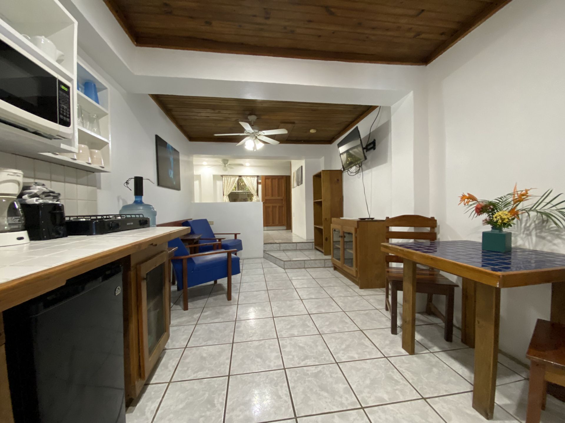 Small apartment interior with kitchen, living area, and dining space. White walls, wood ceiling, tiled floor.