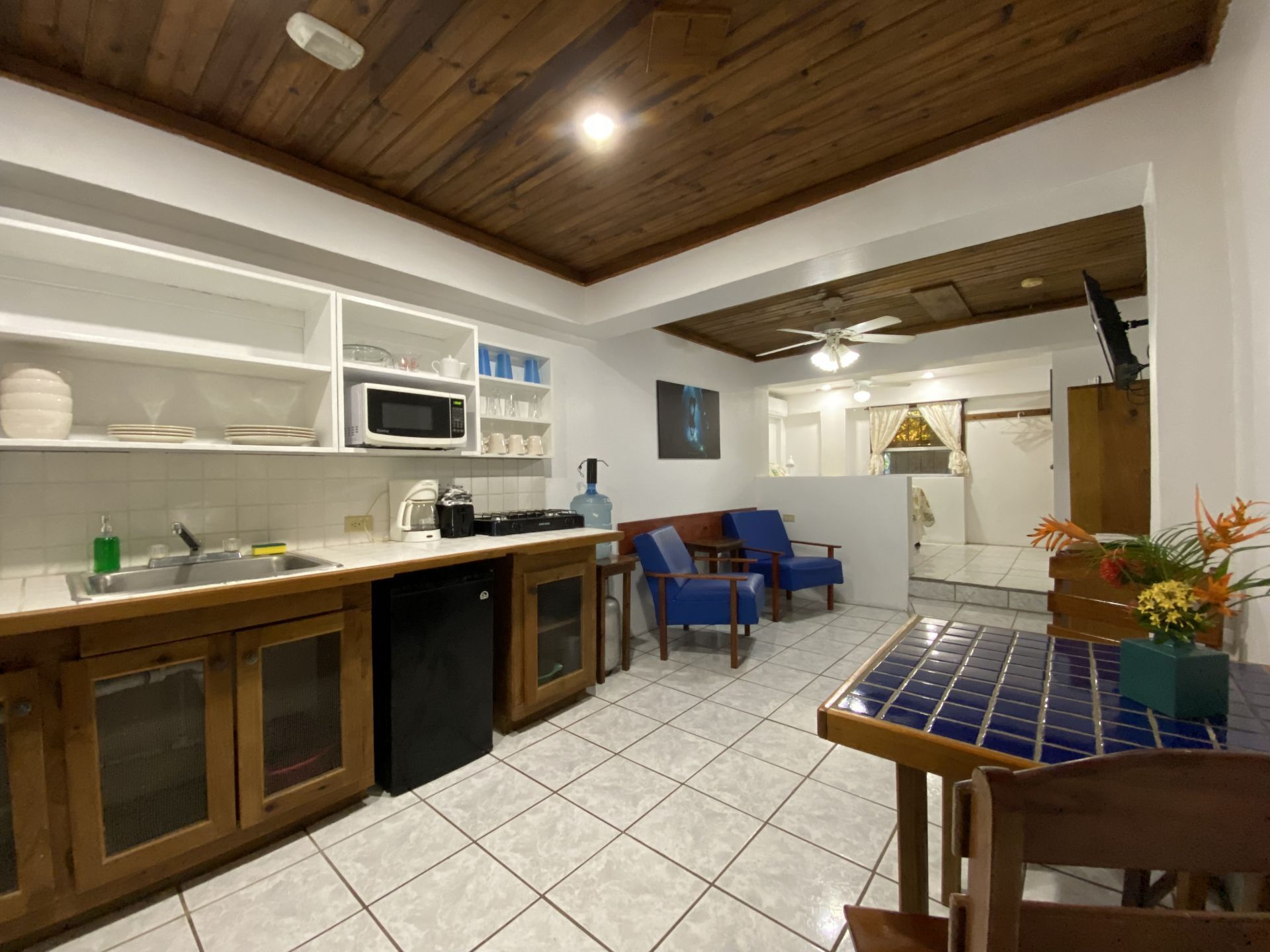 Kitchen and living area. Dark wood cabinets and ceiling, white tile floor, blue armchairs, and tiled dining table.