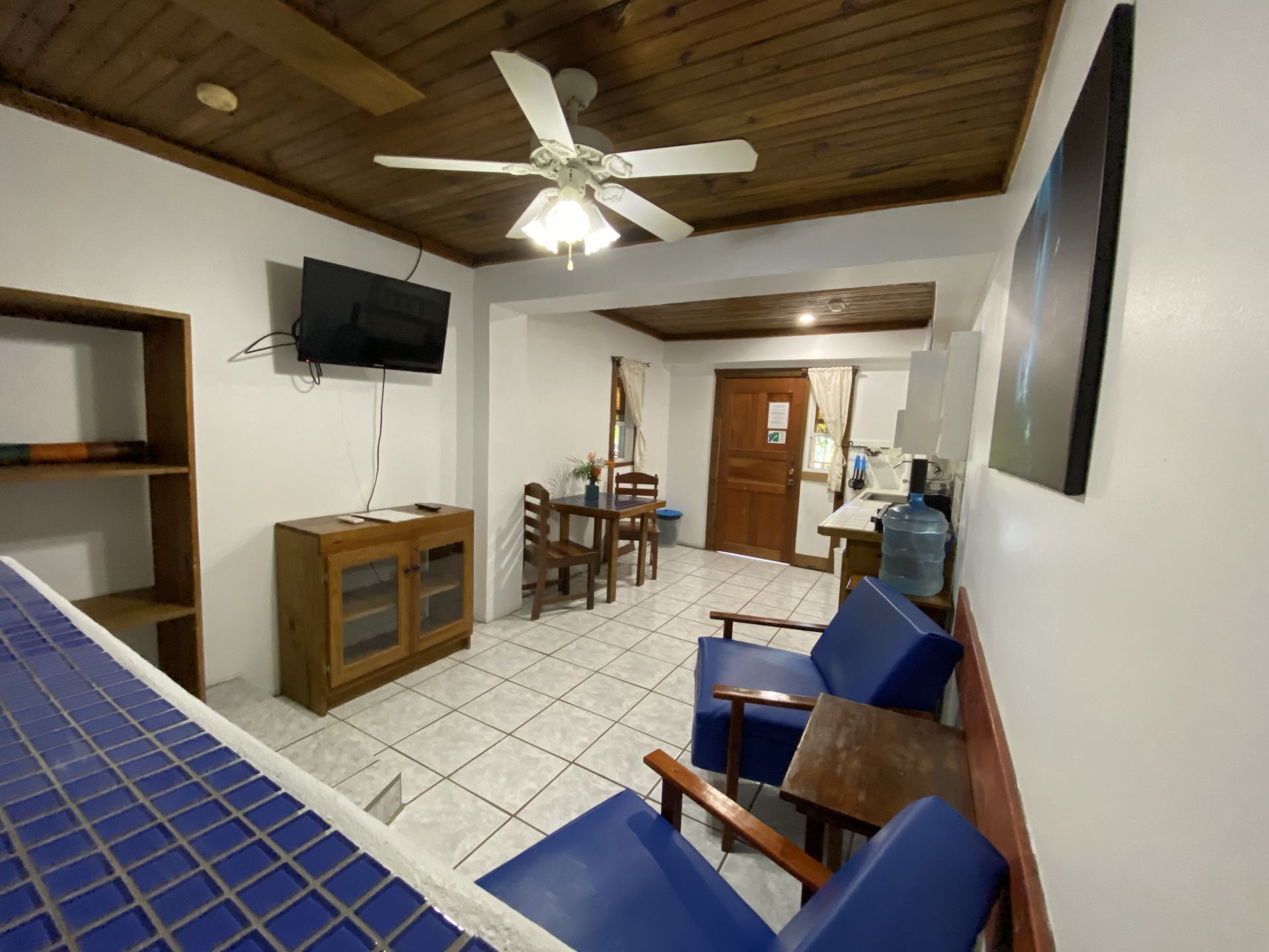 Interior shot of a small apartment with blue tile, wood furniture, and a ceiling fan.