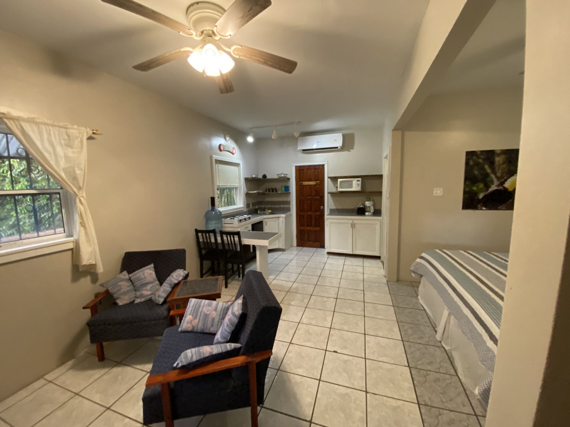 Small studio apartment with seating area, kitchen, and bed. White walls and tile floors.