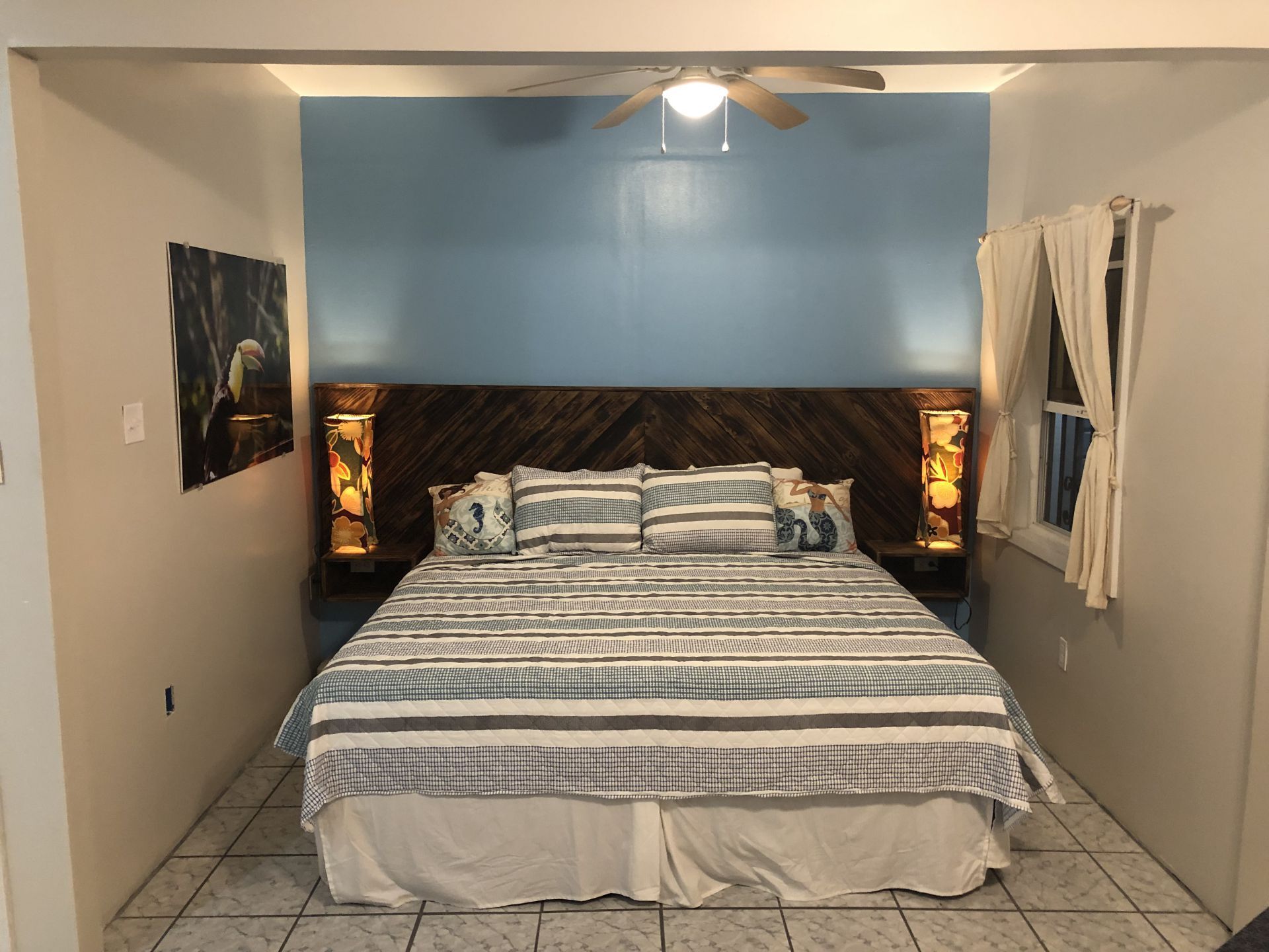 Bedroom with a bed, blue wall, wooden headboard with lights, artwork, and a window.