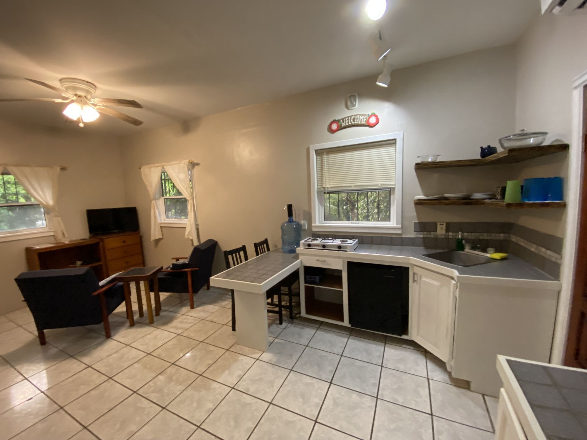 Open-concept kitchen and living area. White tile floor, white cabinets, gray countertops, black armchairs, and a table.