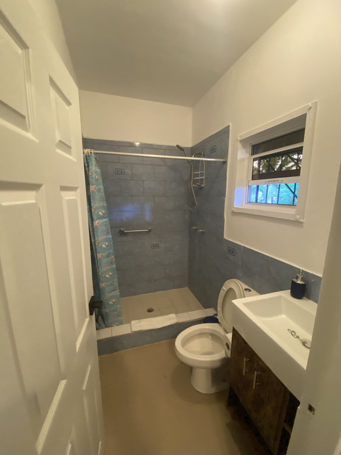 Bathroom with blue tiled shower, white toilet and sink, wooden vanity, and small window.