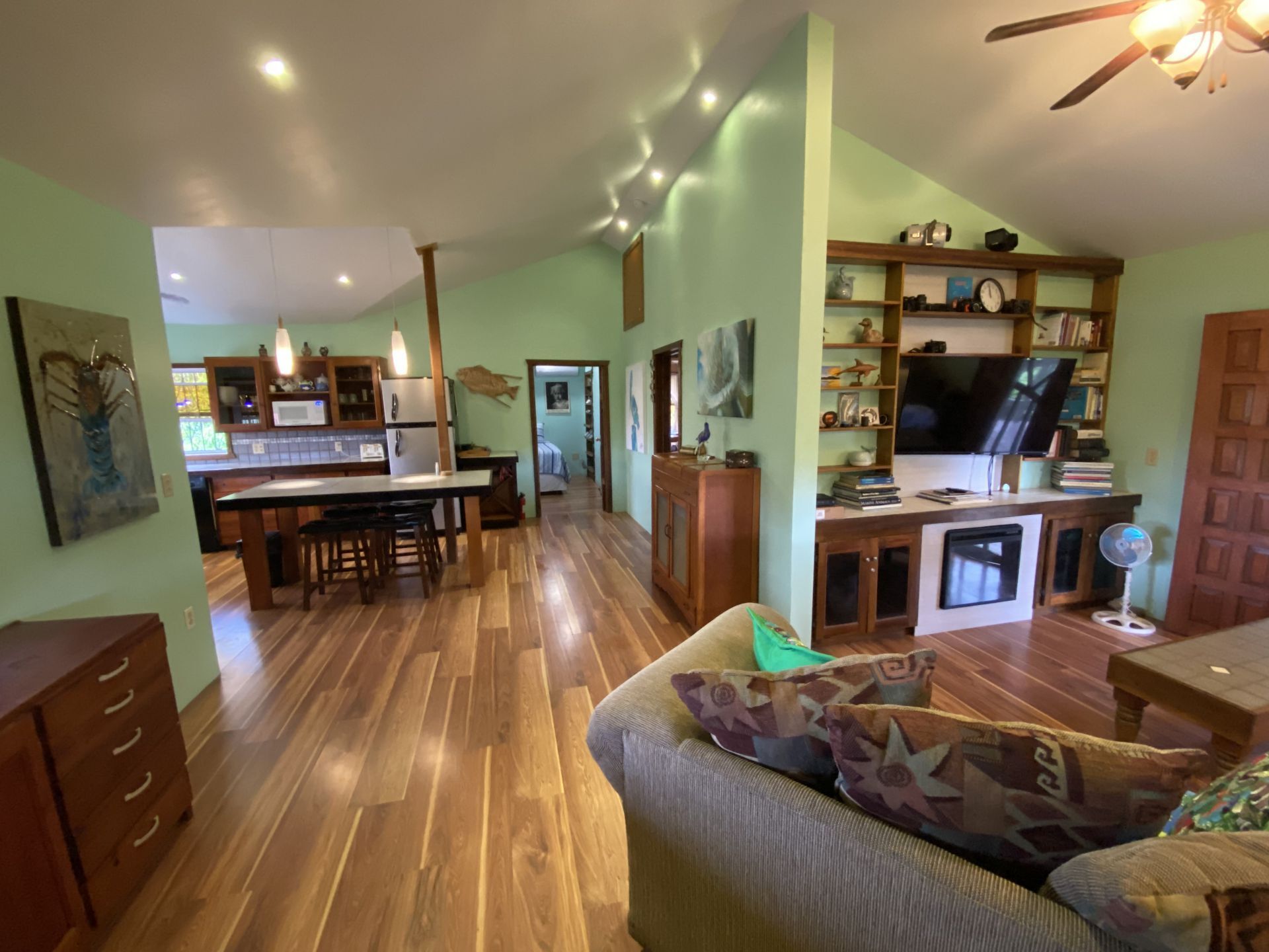 Spacious, light green interior with wooden floors, a living area, and a kitchen; a sofa, TV, and cabinets are present.